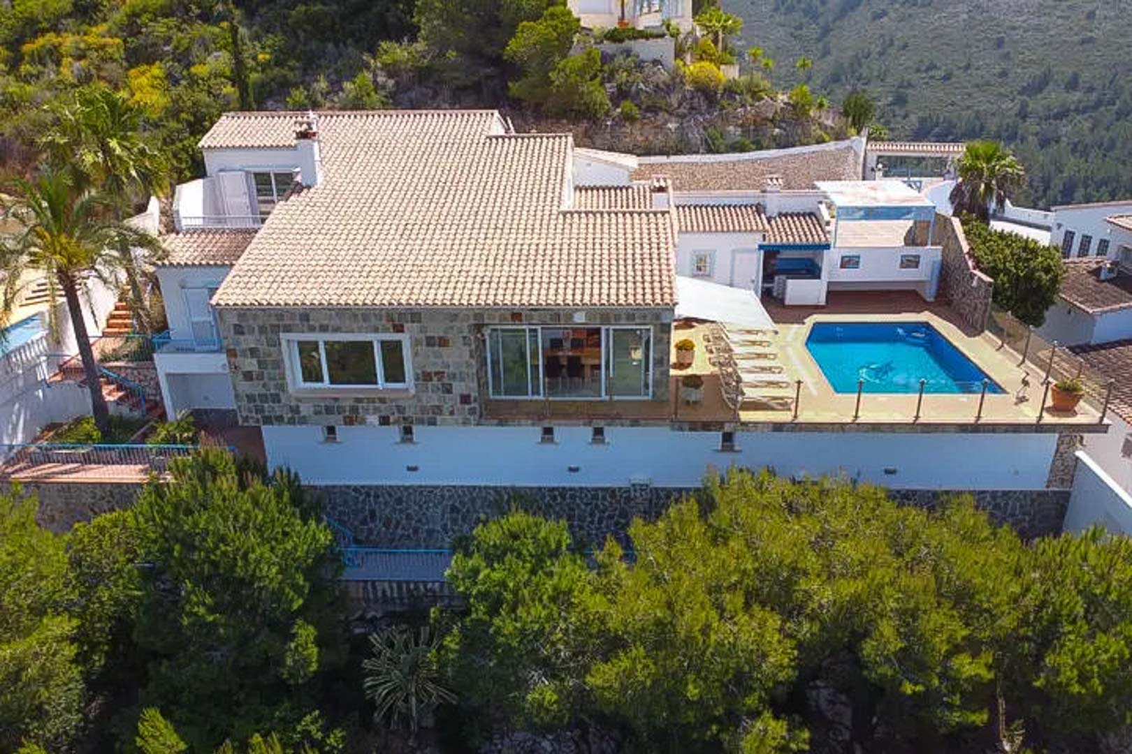Luftaufnahme der Villa mit Natursteinfassade, Ziegeldächern und einer erhöhten Terrasse mit privatem Pool und Sonnenbereich im Grünen.