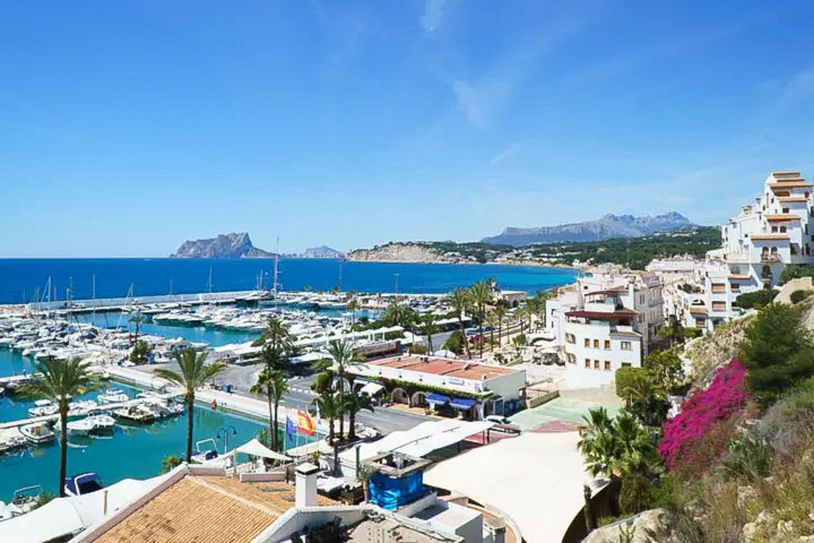 Panoramablick auf den Jachthafen von Moraira mit dem Peñón de Ifach im Hintergrund, traditioneller weißer Architektur und dem türkisblauen Mittelmeer.