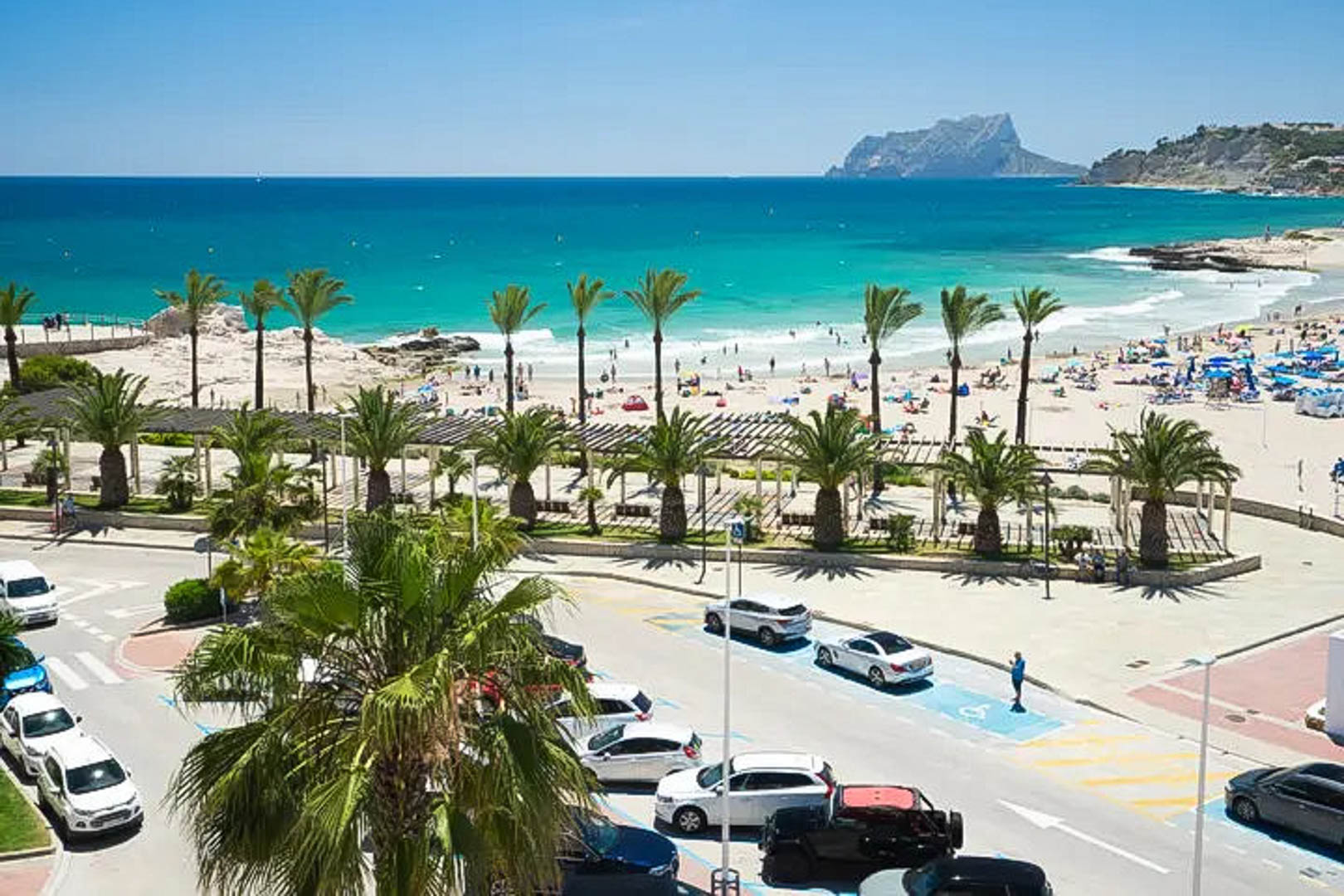 Panoramablick auf den Sandstrand von Moraira mit türkisblauem Wasser, dem Peñón de Ifach im Hintergrund und einer von Palmen gesäumten Promenade.