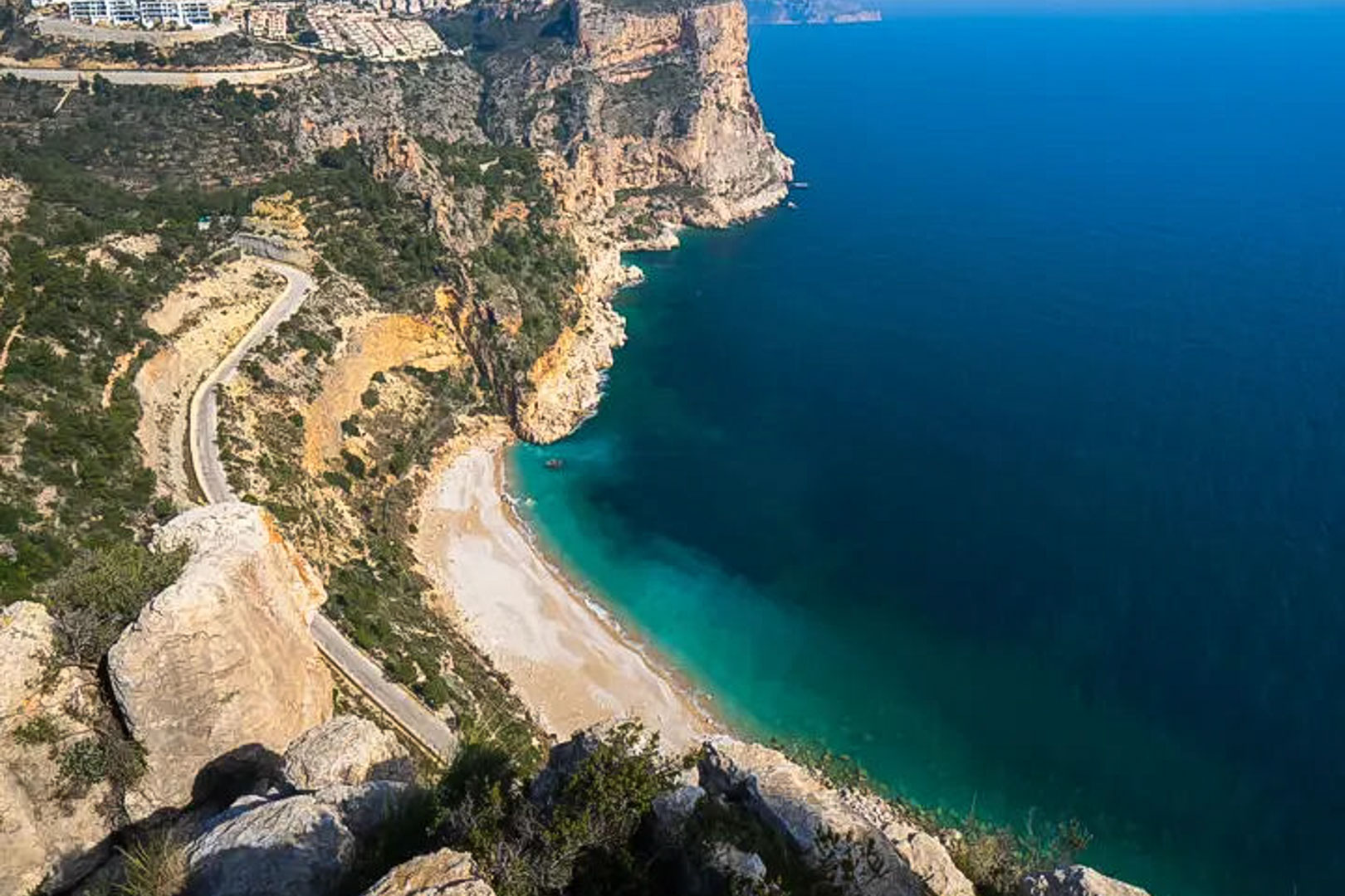 Panoramablick auf die Klippen der Costa Blanca mit einer türkisfarbenen Bucht und einer kurvenreichen Küstenstraße am Mittelmeer.