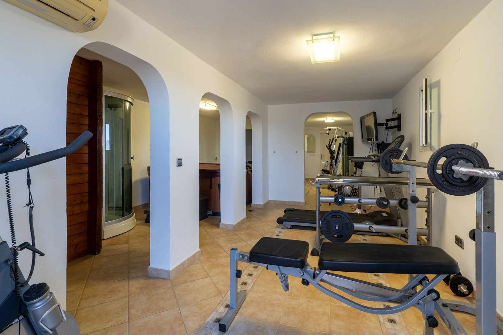 Privater Fitness- und Wellnessbereich mit Kraftgeräten, Laufband und Rundbögen, die zur hauseigenen Sauna, Dampfdusche und zum Whirlpool führen.