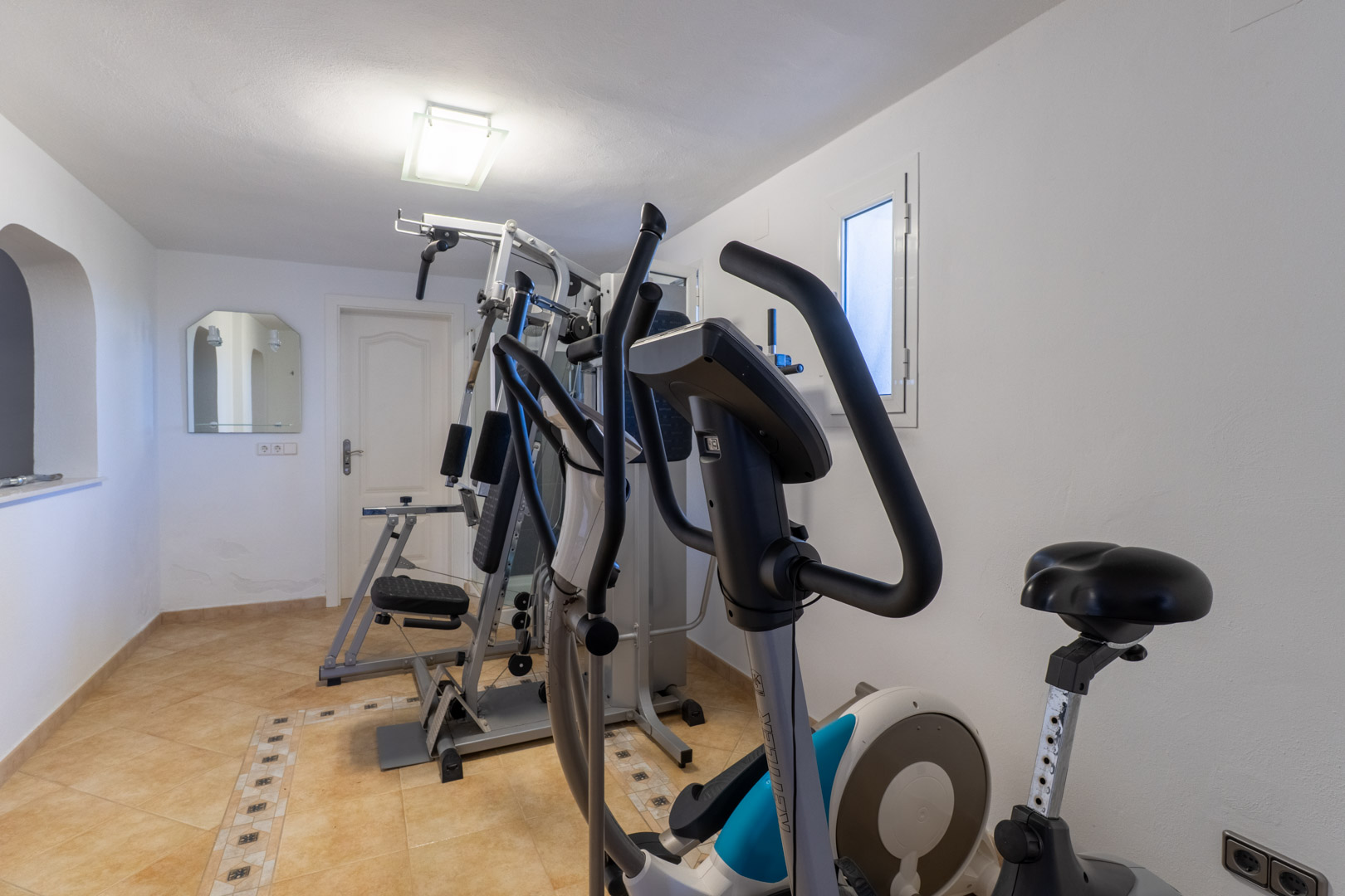 Privater Fitnessraum mit Multikraftstation, Crosstrainer und Heimtrainer auf hellem Fliesenboden mit dekorativer Bordüre.