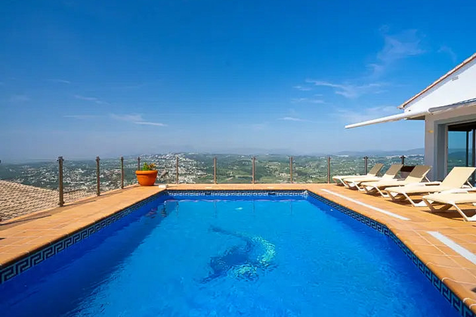 Rechteckiger Pool mit Delfin-Mosaik, Sonnenterrasse mit Liegen und Glasgeländer, das einen weiten Panoramablick auf das Tal und die Küste bietet.