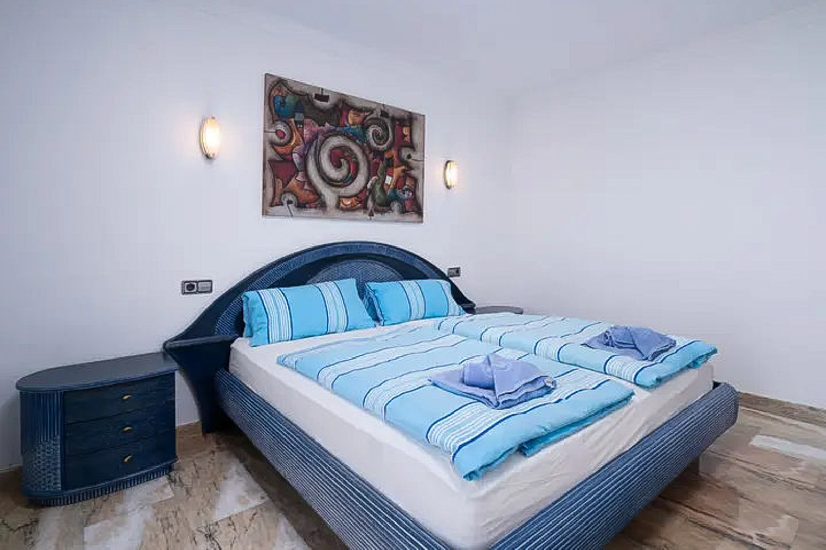 Schlafzimmer mit blauem Korbkopfteil und passendem Nachttisch. Polierter Marmorboden, Wandleuchten und ein abstraktes Gemälde ergänzen den Raum.