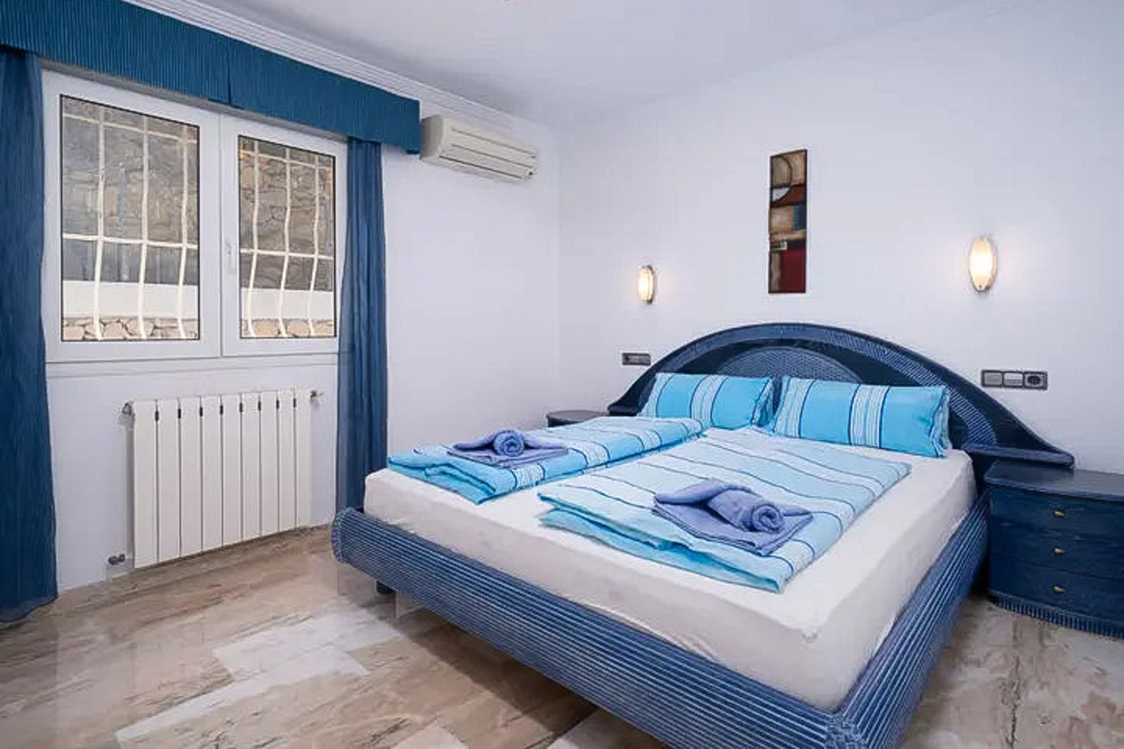 Schlafzimmer mit poliertem Marmorboden, Doppelbett in Blautönen, Klimaanlage, Heizkörper und gesichertem Fenster mit passenden Vorhängen.