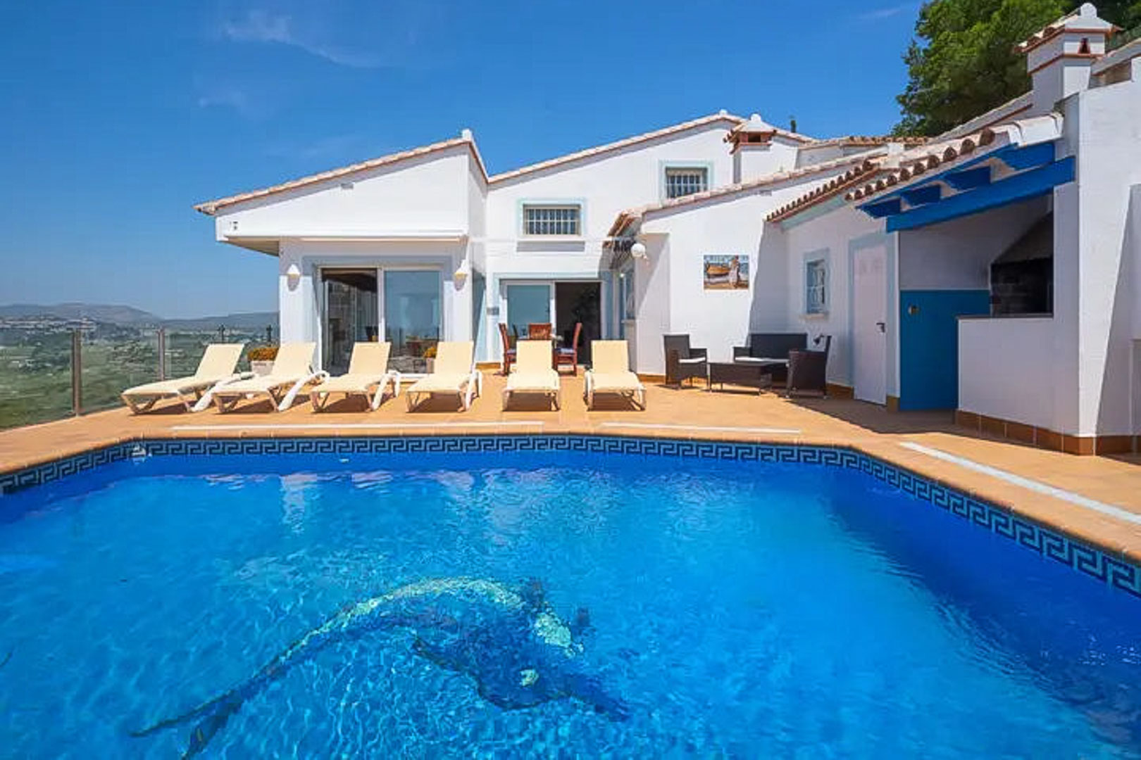 Terrasse mit Pool und Delfinmosaik, Sonnenliegen und Grillbereich unter blauen Holzbalken, mit weitem Blick über die umliegende Landschaft.
