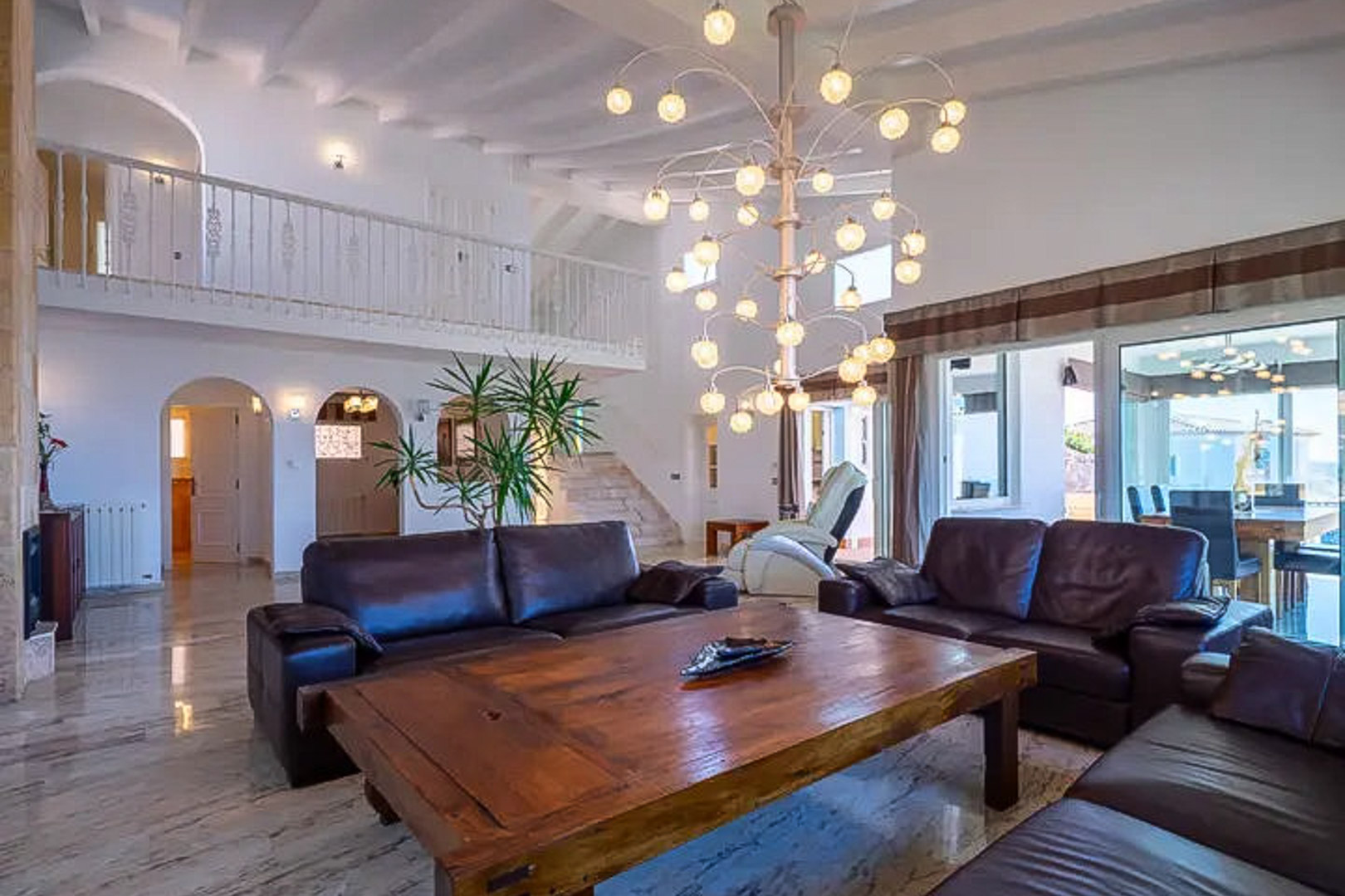 Wohnzimmer mit doppelter Deckenhöhe, dunklen Ledersofas, Massivholz-Couchtisch und Designer-Kronleuchter. Marmorböden und eine weiße Galerie prägen den offenen Raum.