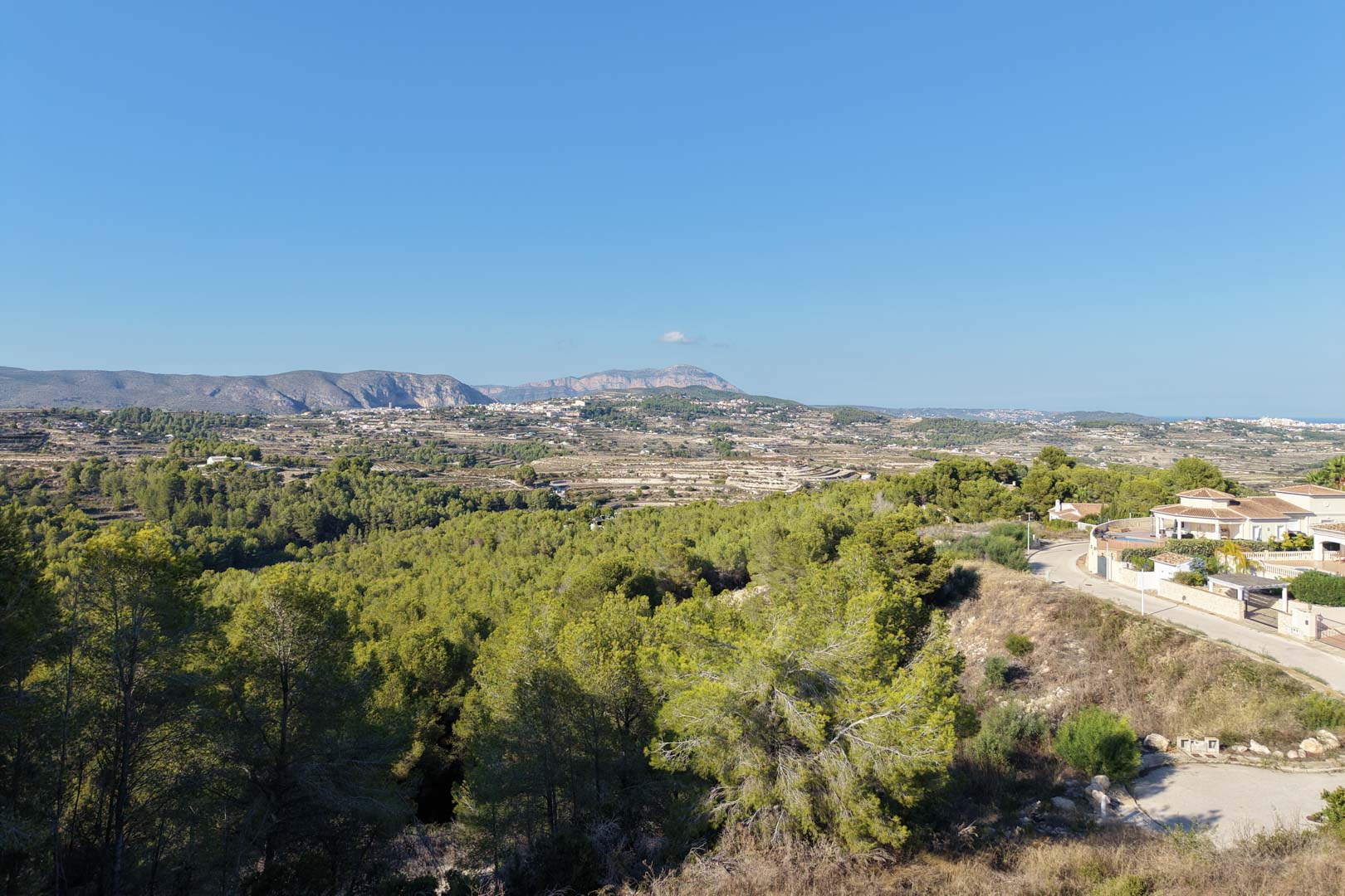 Erhöhter Panoramablick über das Tal und die fernen Berge, umgeben von mediterranen Pinienwäldern und angrenzenden Wohngebieten.