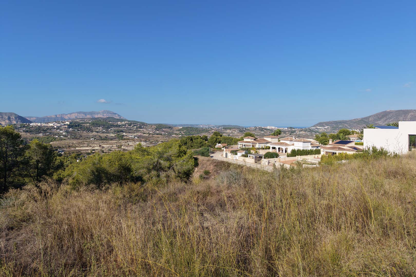 Erhöhtes Baugrundstück mit natürlicher Vegetation und Panoramablick auf das Tal, die umliegenden Berge und das Mittelmeer am Horizont.