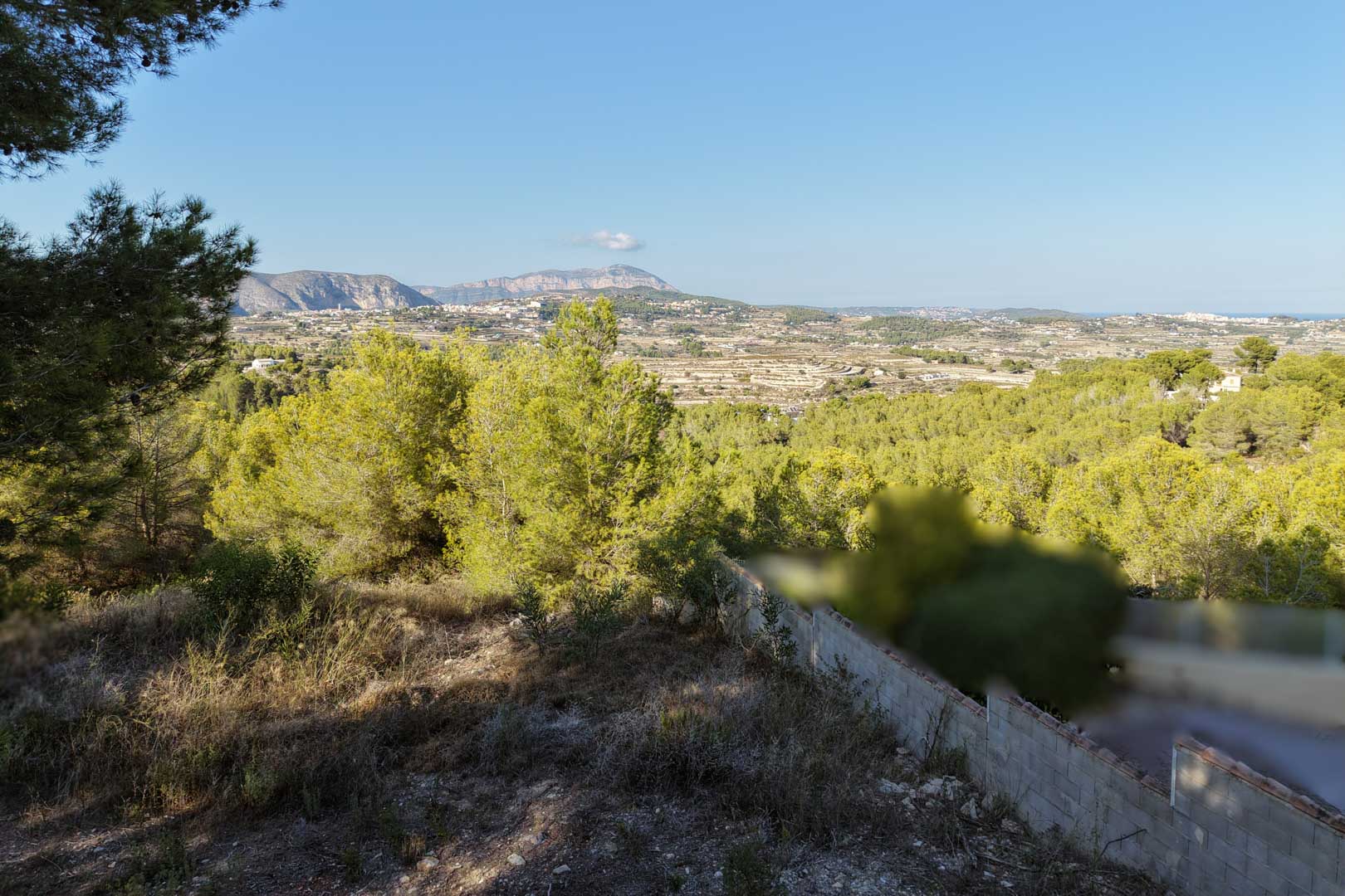 Erhöhter Panoramablick vom Grundstück über das Tal von Moraira mit einheimischen Pinienwäldern und fernen Bergketten unter klarem blauem Himmel.
