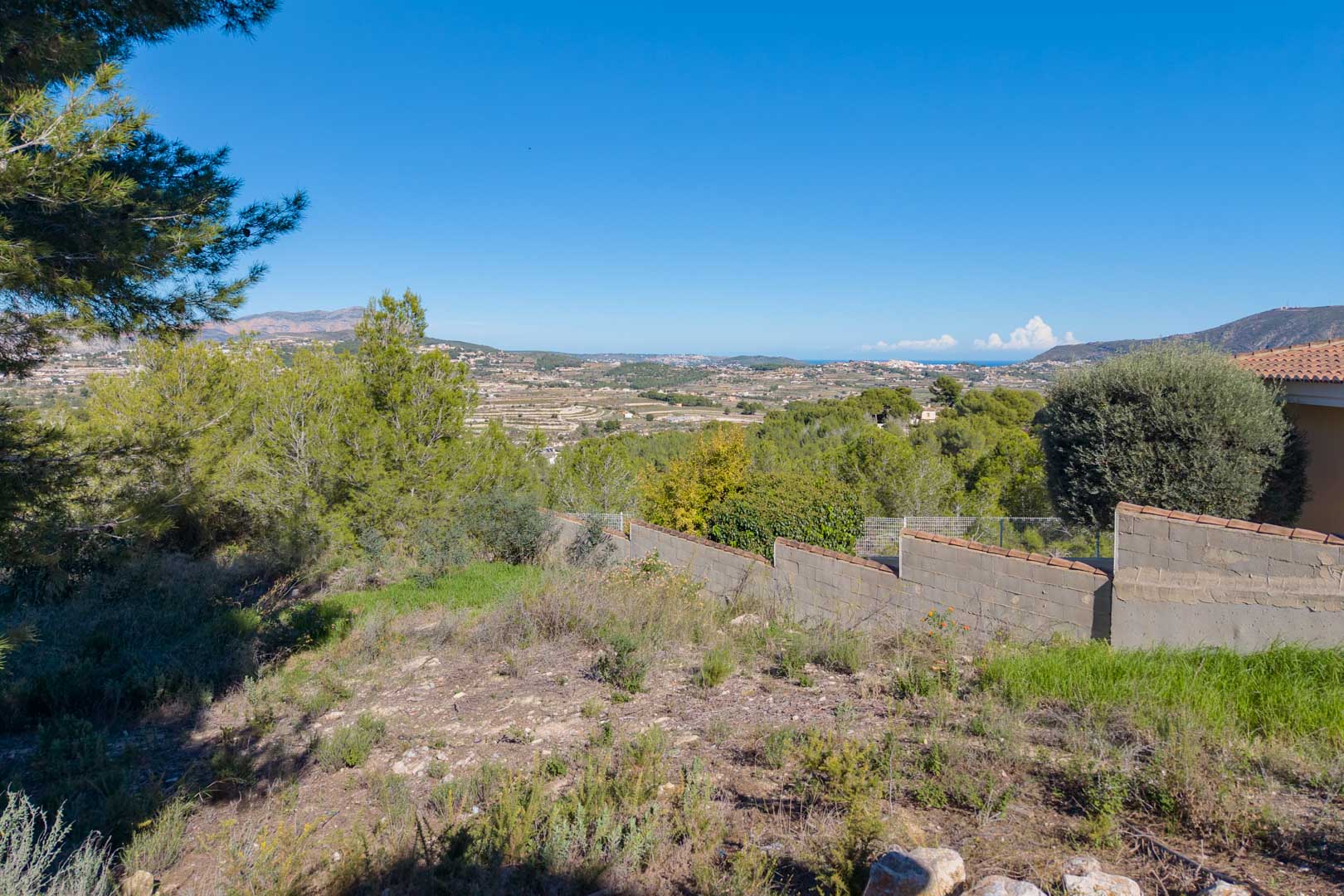 Erhöhtes Grundstück mit Panoramablick auf das Tal und das Meer. Das Gelände umfasst natürliche Vegetation und eine Begrenzungsmauer mit Ziegelabdeckung.