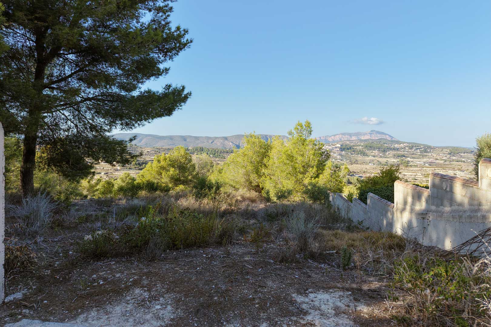 Erhöhtes Grundstück mit Panoramablick auf das Tal und die Berge. Das Gelände umfasst natürliche Vegetation, Pinien und eine abgestufte Begrenzungsmauer.