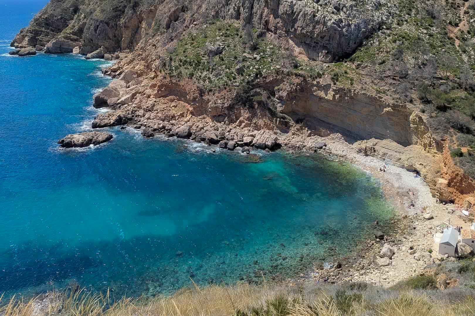Luftaufnahme einer Mittelmeerbucht mit türkisfarbenem Wasser, schroffen Kalksteinfelsen und einem kleinen Kieselstrand an der Küste von Moraira.