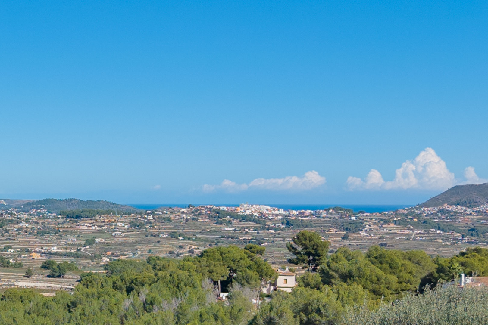 Panoramablick vom Grundstück in Benimeit über terrassierte Hänge, mediterrane Pinienwälder und das ferne Meer unter klarem blauem Himmel.