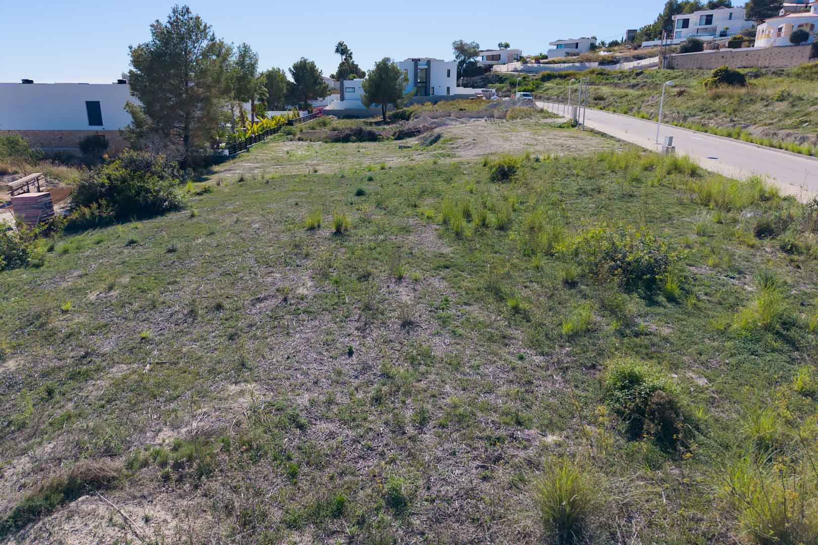Ebenes Stadtgrundstück in El Tesoro, Moraira, mit asphaltiertem Straßenzugang und Blick auf moderne Villen. Das Gelände bietet eine offene Aussicht auf das Tal.