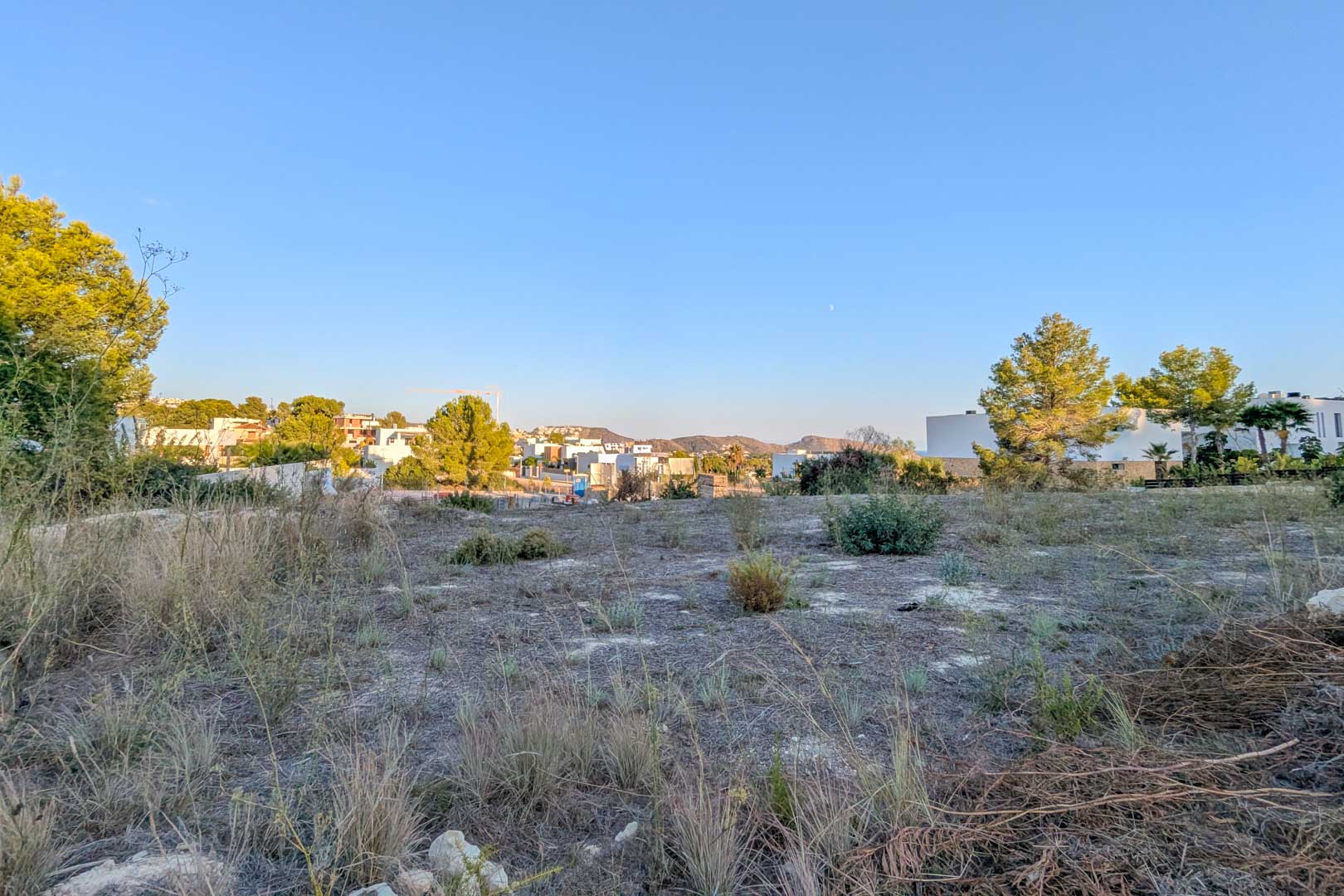 Panoramablick auf das flache Baugrundstück in El Tesoro, umgeben von mediterraner Vegetation und modernen weißen Villen vor einer Hügelkulisse.