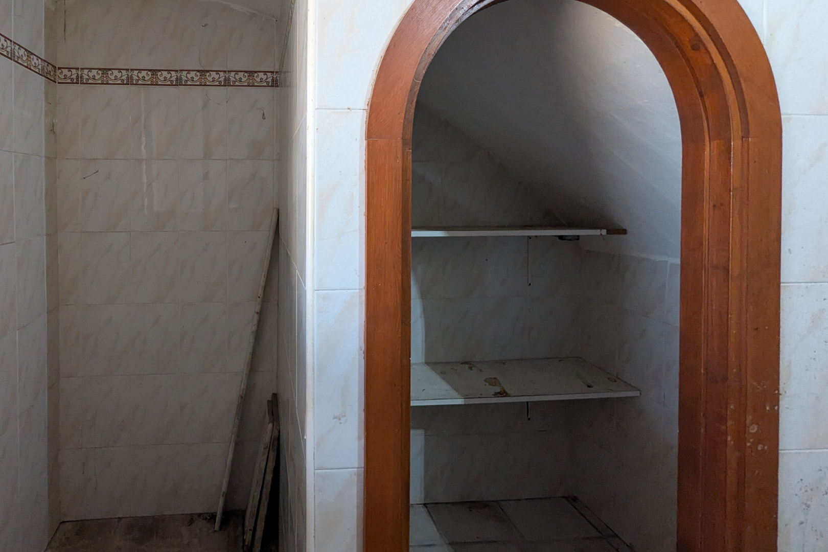 Abstellnische unter der Treppe mit Holzrundbogen, weiß gefliesten Wänden und praktischen Regalböden für zusätzlichen Stauraum.