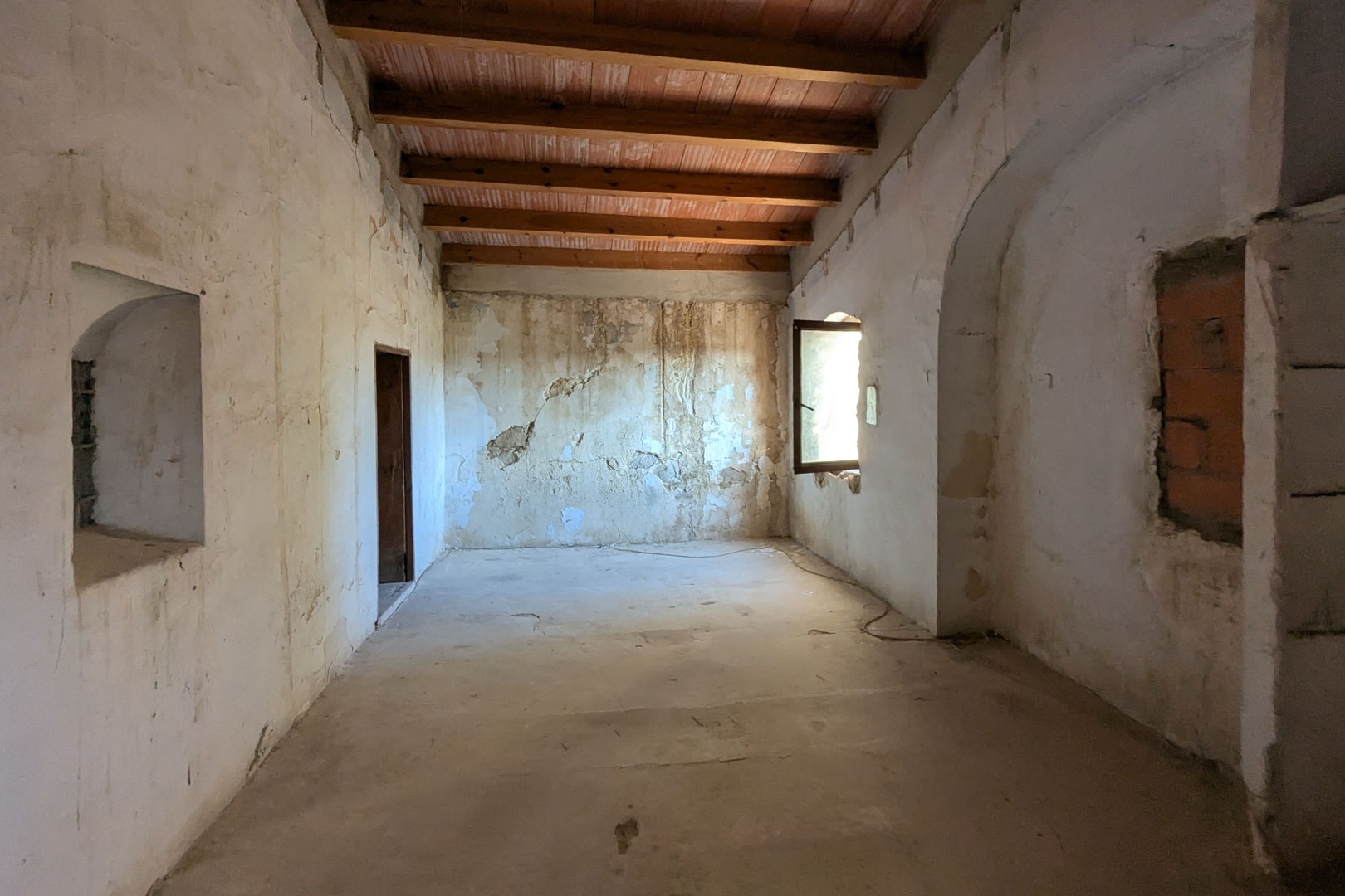 Großzügiger, offener Raum mit hohen Decken aus Holzbalken und Terrakotta-Fliesen. Die dicken Steinwände verfügen über originale Nischen und ein Fenster mit natürlichem Lichteinfall.