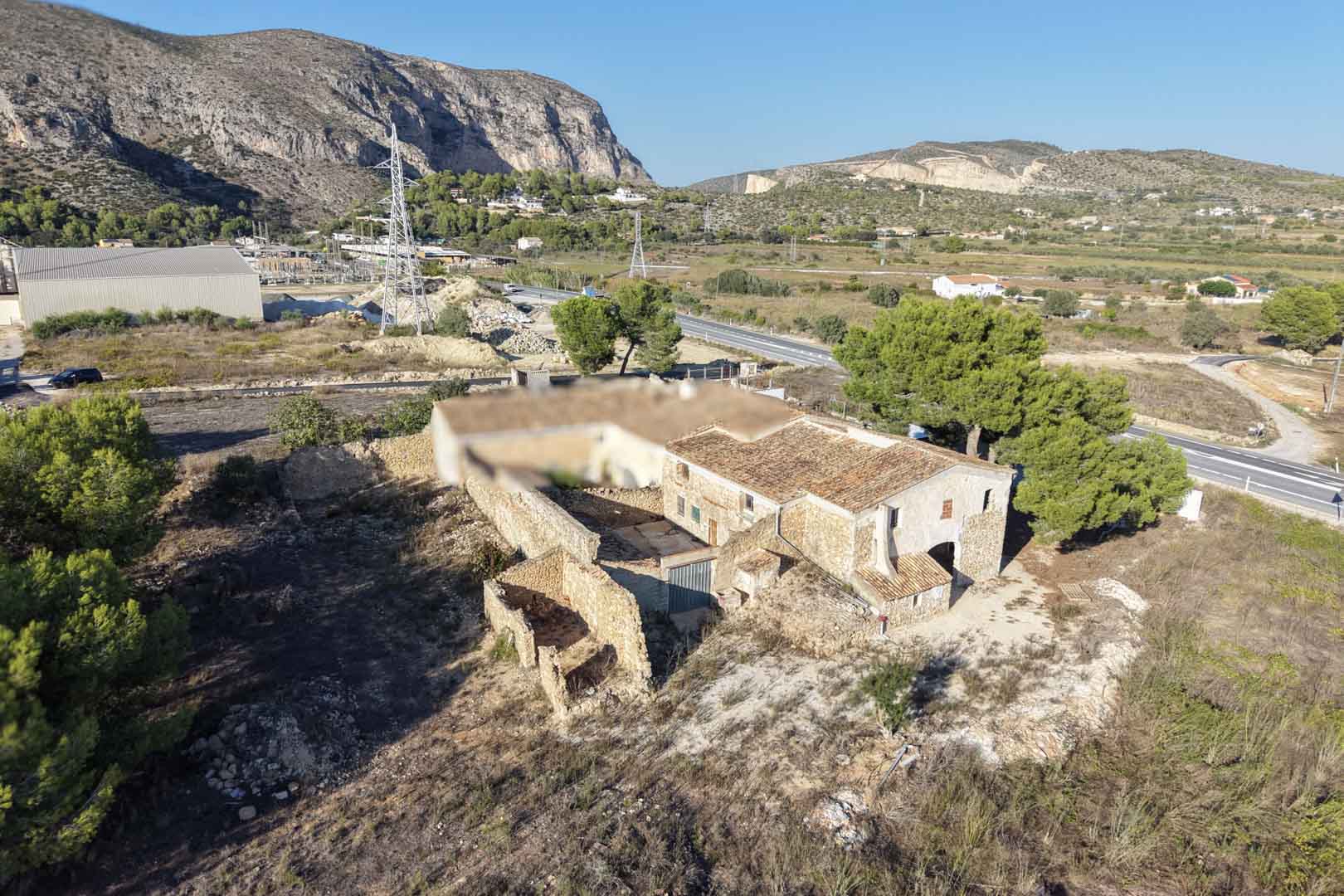 Luftaufnahme des traditionellen Natursteinhauses mit Innenhof und Nebengebäuden auf einem weitläufigen rustikalen Grundstück mit Blick auf die Berge von Teulada.