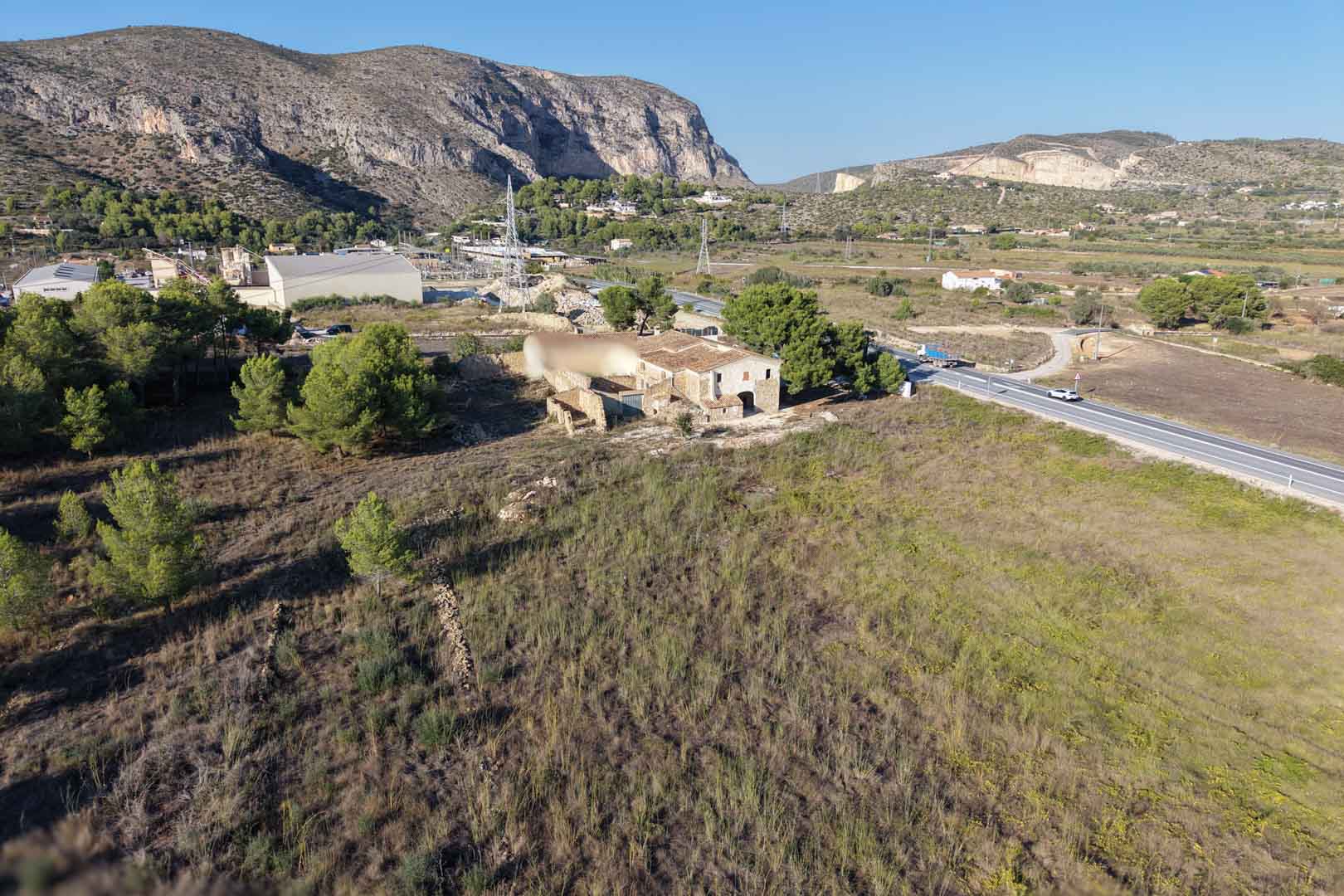 Luftaufnahme des traditionellen Steinhauses auf einem weitläufigen, flachen Grundstück in Teulada, umgeben von Bergen und mediterraner Landschaft.