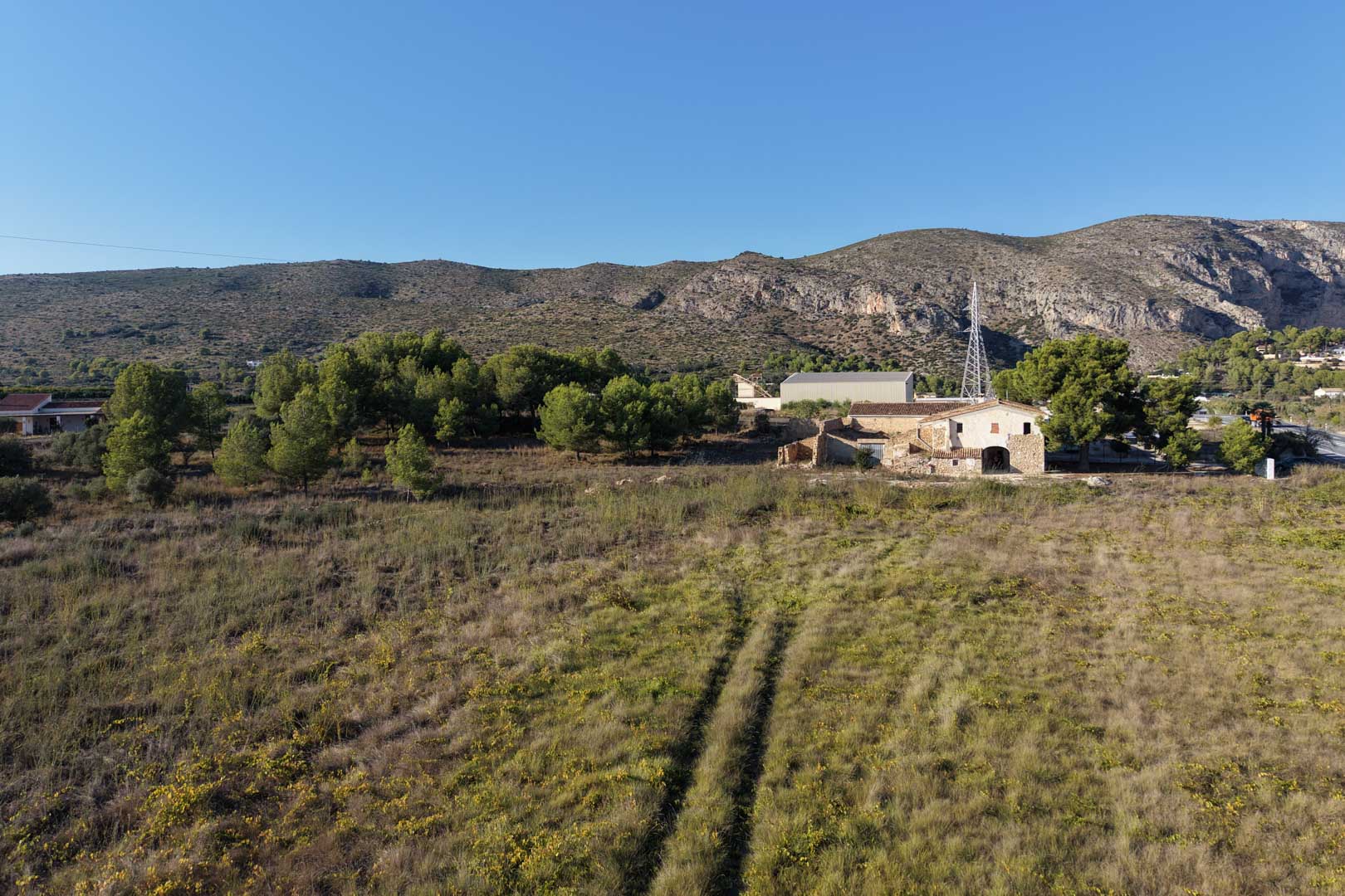 Luftaufnahme des traditionellen Steinhauses und seines weitläufigen, flachen Grundstücks, umgeben von mediterraner Vegetation und den Bergen von Teulada im Hintergrund.