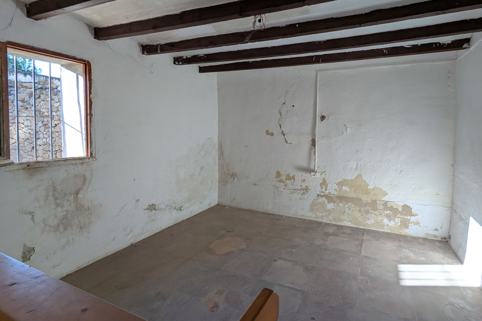 Rustikaler Innenraum mit originalen dunklen Holzbalken, kalkverputzten Wänden und traditionellem Fliesenboden. Ein vergittertes Fenster lässt natürliches Licht in den geschichtsträchtigen Raum.