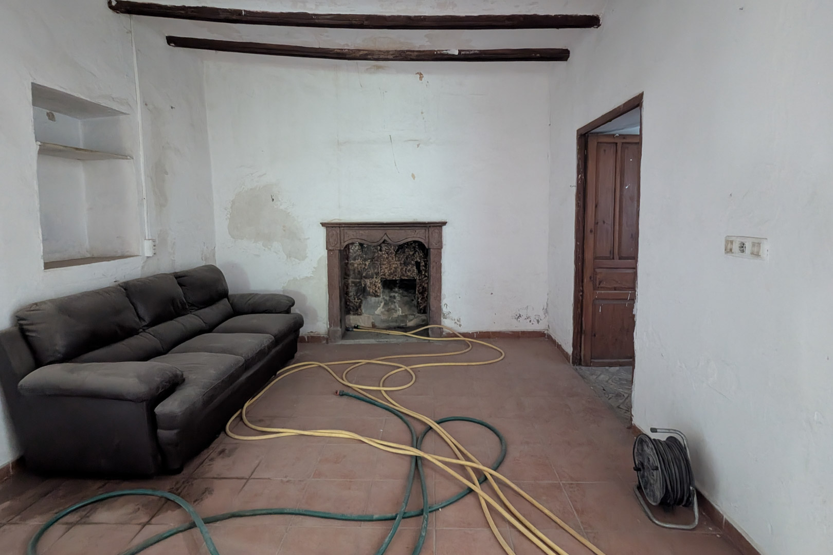 Traditionelles Wohnzimmer mit sichtbaren Holzbalken, Terrakotta-Fliesenboden und einem originalen Kamin mit geschnitzter Steineinfassung.