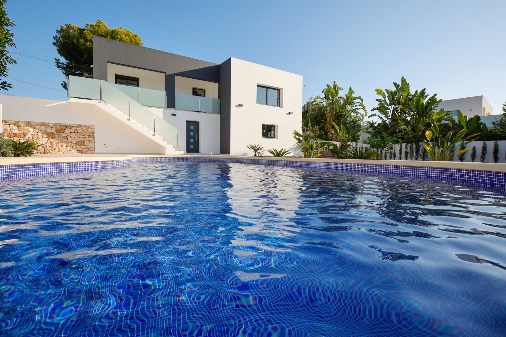 Außenansicht der Villa mit blauem Mosaikpool, minimalistischer Fassade in Weiß und Grau, Natursteinmauern und Terrassen mit Glasgeländern.