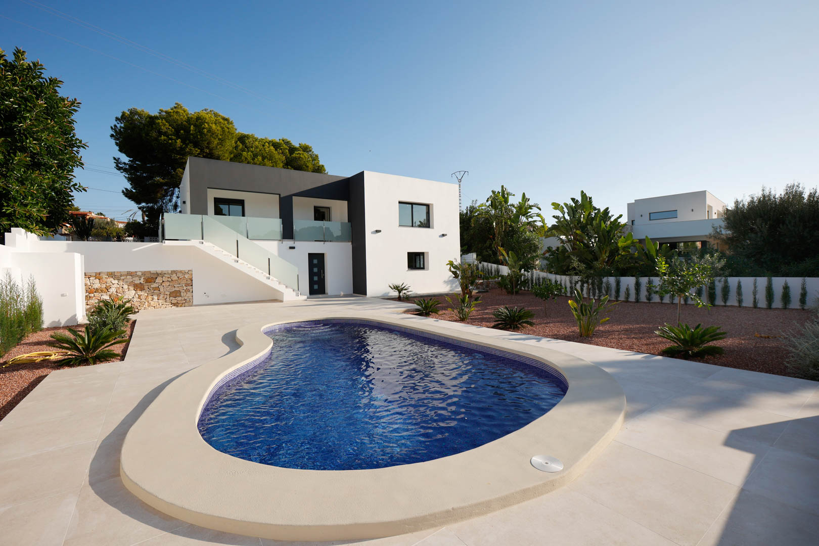 Außenansicht mit geschwungenem Pool, Feinsteinzeug-Terrasse und Garten mit rotem Marmorkies. Moderne zweifarbige Fassade mit Außentreppe und Glasgeländern.