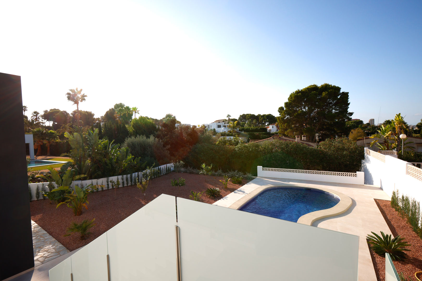 Blick von der oberen Terrasse mit Glasgeländer auf den geschwungenen Pool, den Garten mit rotem Marmorkies und die mediterrane Umgebung.