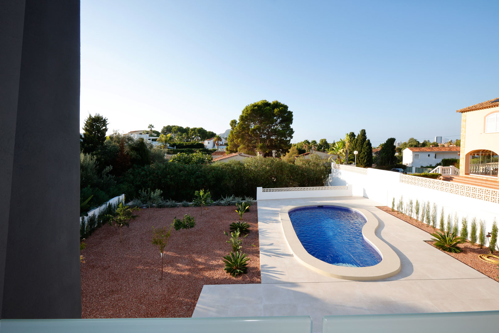 Erhöhter Blick auf den Garten mit nierenförmigem Pool, weißer Fliesenterrasse und einer Fläche aus rotem Marmorkies mit mediterraner Bepflanzung.