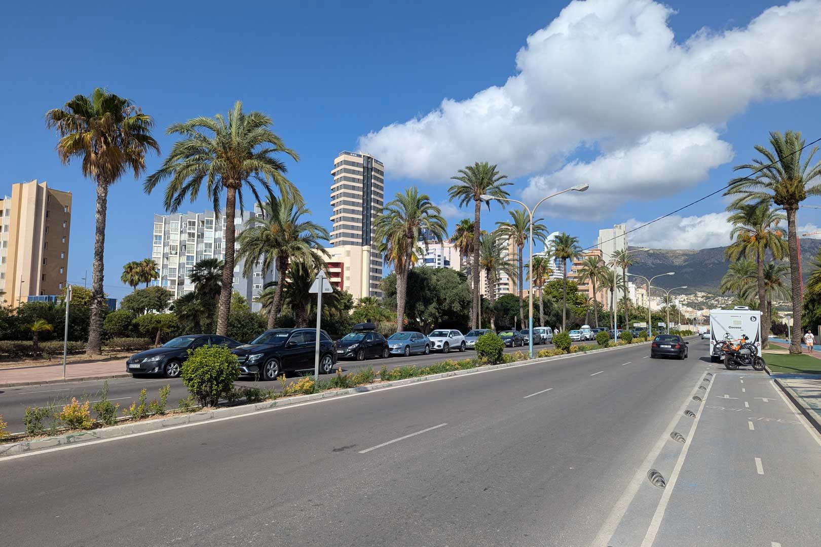 Küstenstraße mit Radweg, Palmen und Hochhäusern vor einer Bergkulisse unter strahlend blauem Himmel.