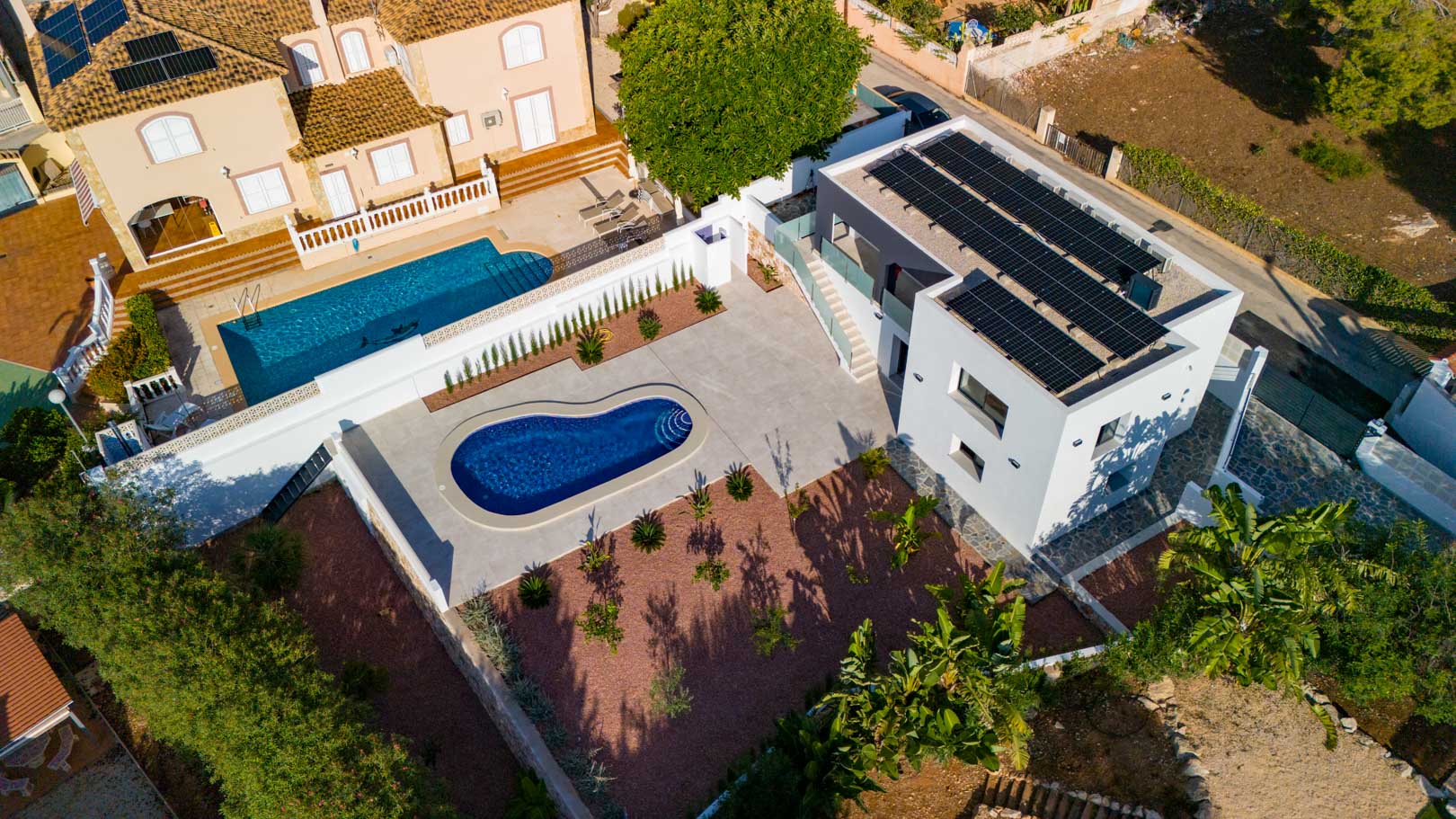 Luftaufnahme der modernen Villa mit Photovoltaikanlage, nierenförmigem Pool und Garten mit rotem Marmorkies. Mit Glasgeländern und Natursteinmauern gestaltet.