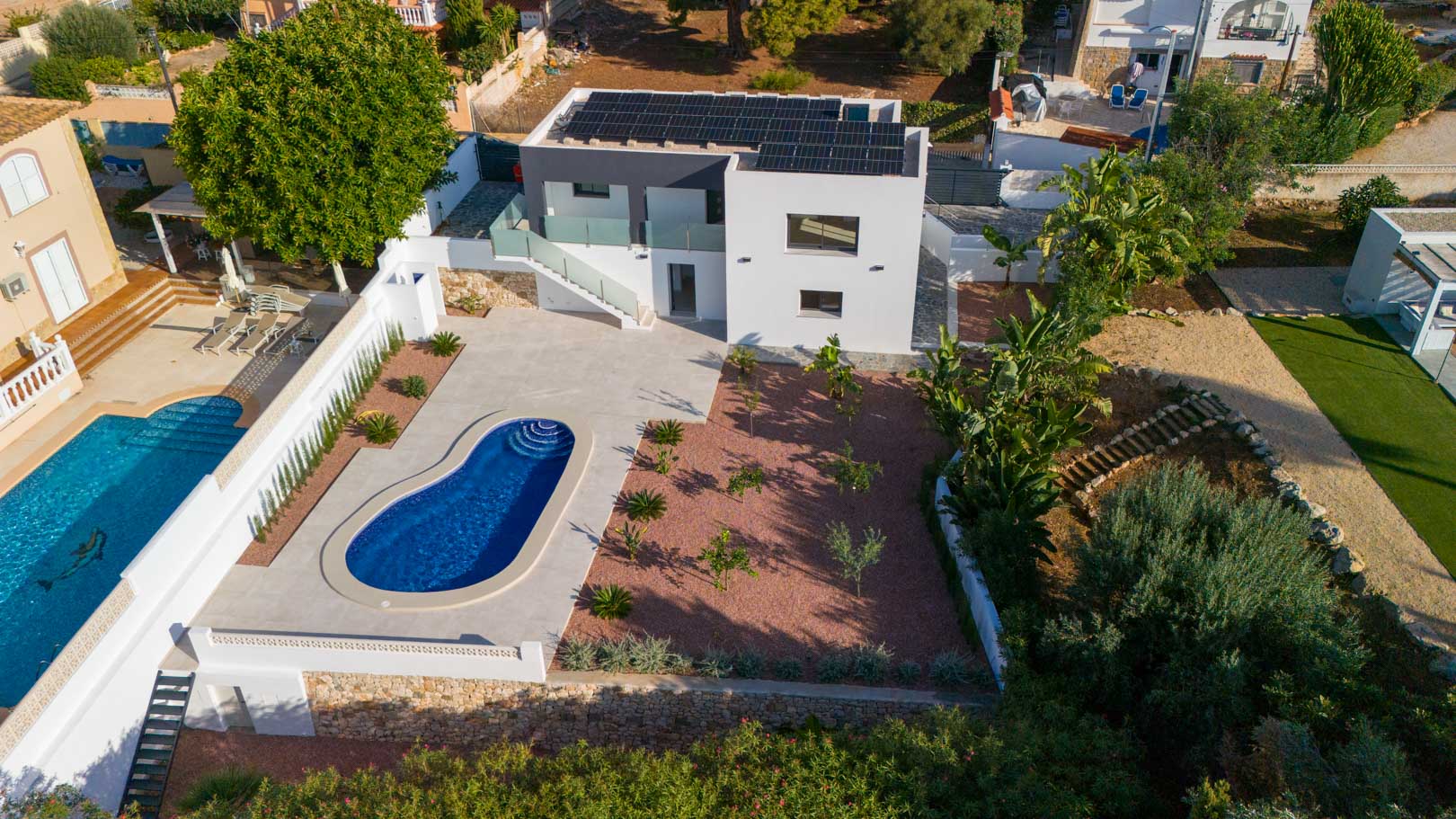 Luftaufnahme der modernen Villa mit Photovoltaikanlage, nierenförmigem Pool und Garten mit rotem Marmorkies und Natursteinmauern.