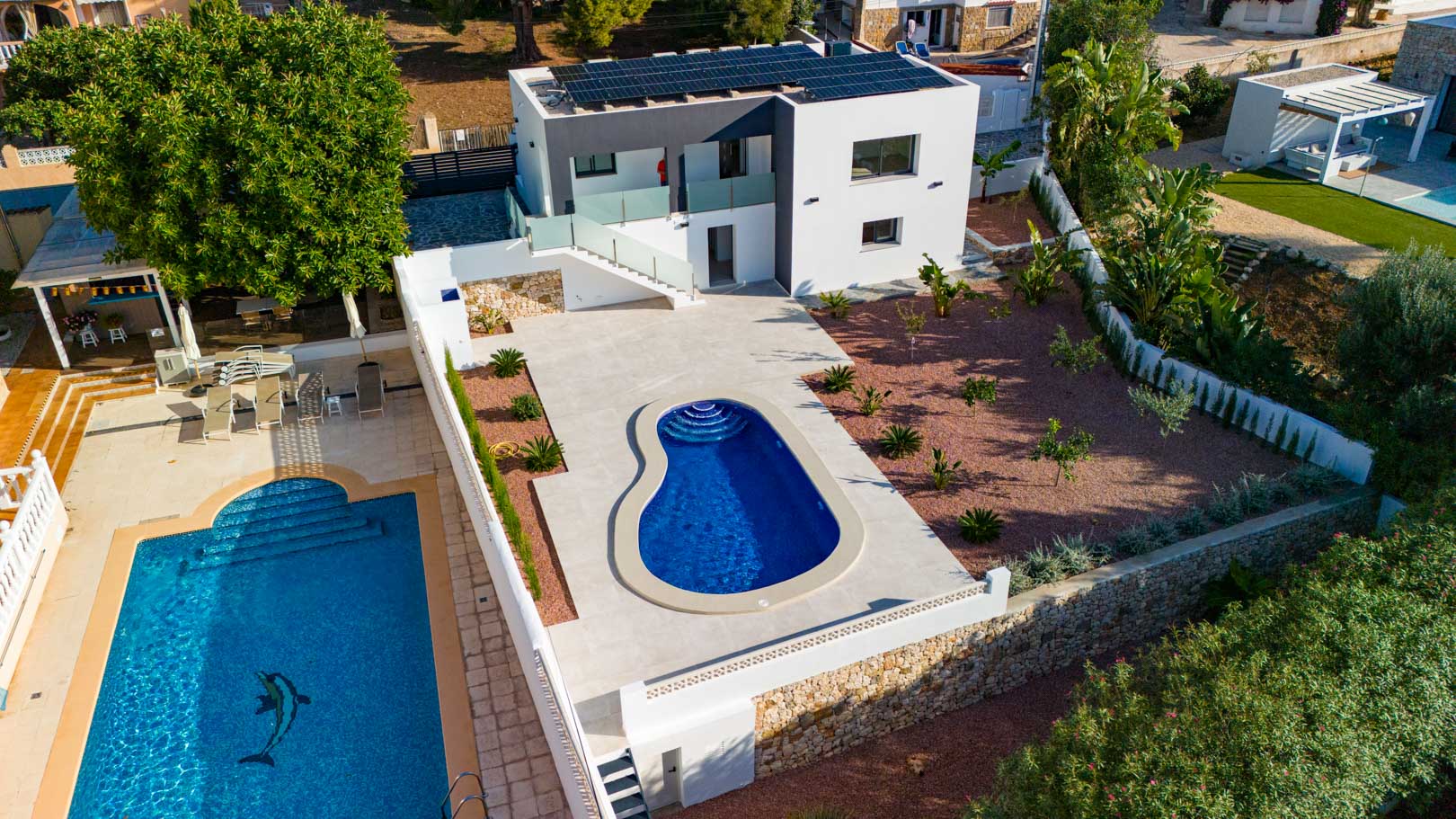 Luftaufnahme der modernen Villa mit Solarmodulen, organisch geformtem Pool, rotem Kiesgarten und Natursteinmauern. Glasgeländer unterstreichen die zeitgenössische Architektur.
