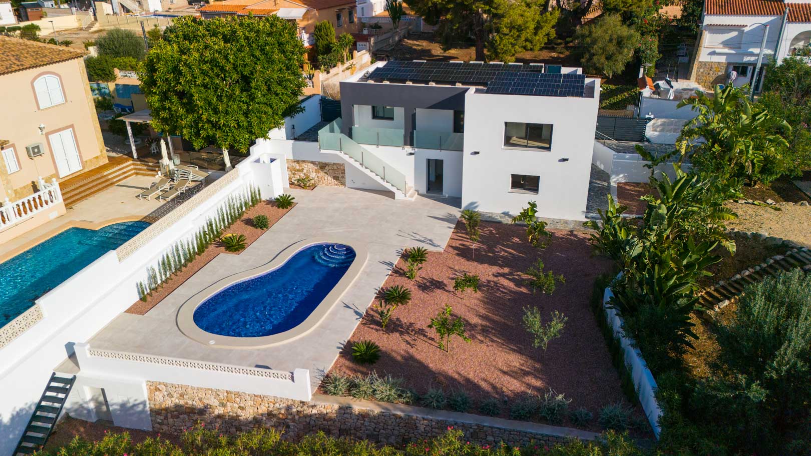 Luftaufnahme der Villa mit nierenförmigem Pool, Garten mit rotem Marmorkies und Photovoltaikanlage. Glasgeländer und Natursteinmauern prägen das Design.
