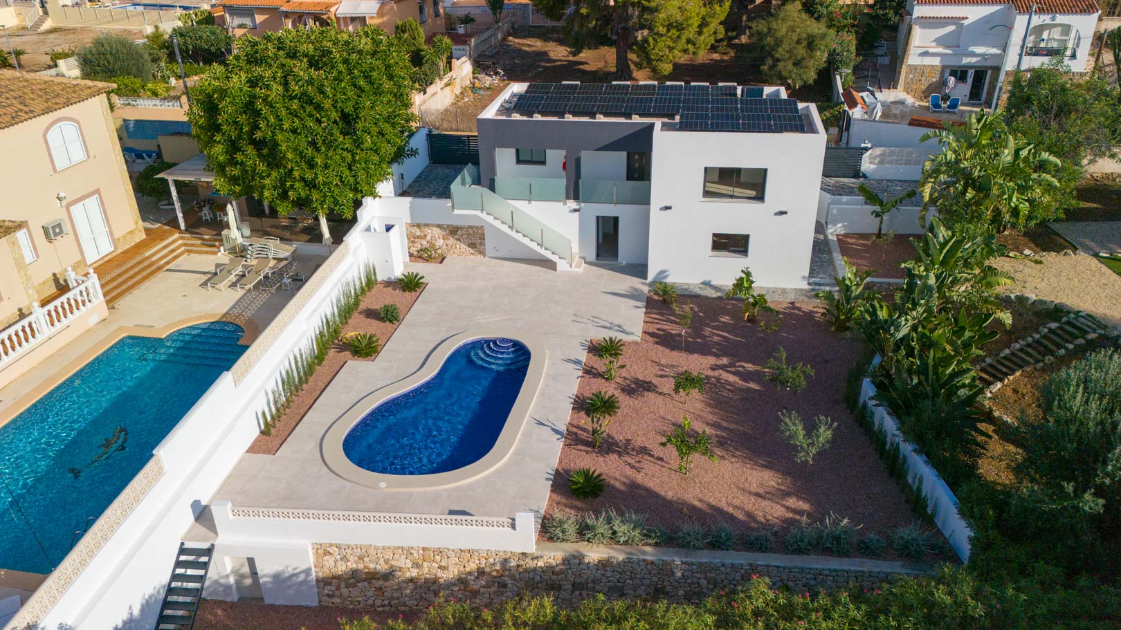 Luftaufnahme der Villa mit nierenförmigem Pool, rotem Kiesgarten und Photovoltaikanlage auf dem Dach. Glasgeländer und Natursteinmauern prägen das moderne Außendesign.