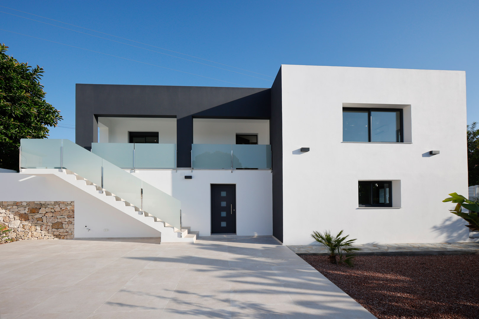 Minimalistische Fassade in Weiß und Grau mit Außentreppe, Glasgeländern, Technal-Aluminiumfenstern und einer dekorativen Natursteinmauer.