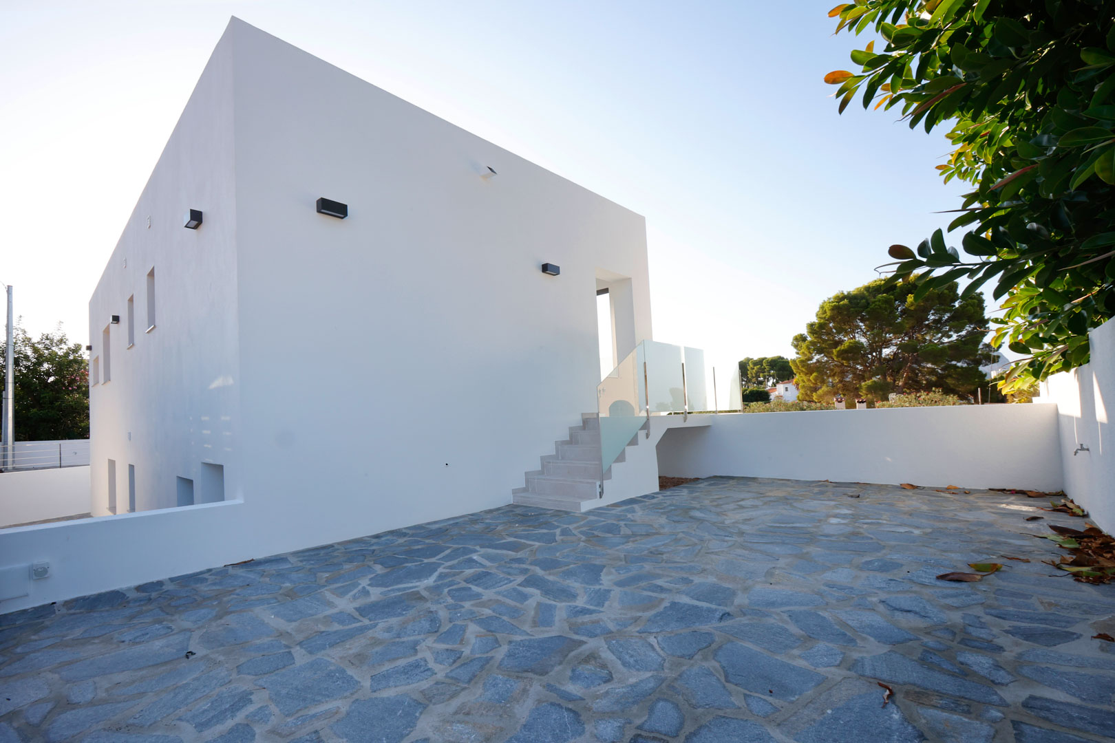 Minimalistische weiße Fassade mit einer großzügigen Terrasse aus unregelmäßigem Naturstein und Außentreppe mit Glas- und Edelstahlgeländer.