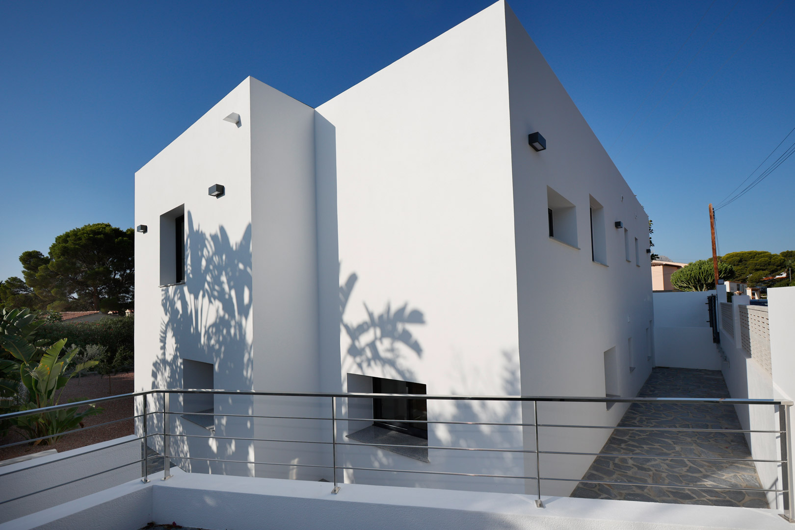 Minimalistische weiße Fassade mit klaren geometrischen Linien, Edelstahlgeländern und Natursteinpflaster. Schattenwürfe betonen das moderne architektonische Design.