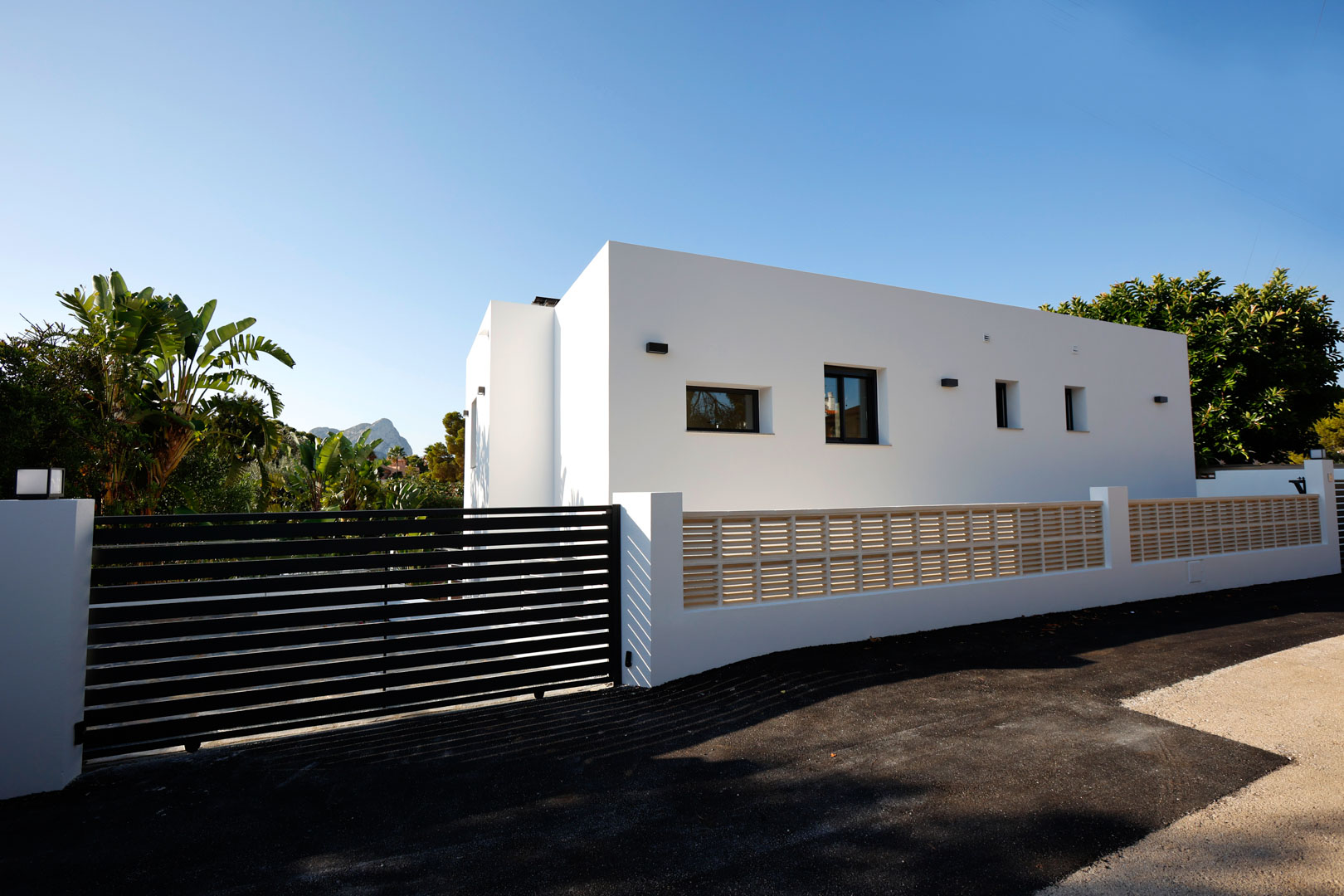 Minimalistische weiße Fassade mit schwarzem Lamellen-Automatiktor, Umfassungsmauer mit dekorativem Gitter und gepflasterter Zufahrt vor Bergkulisse.