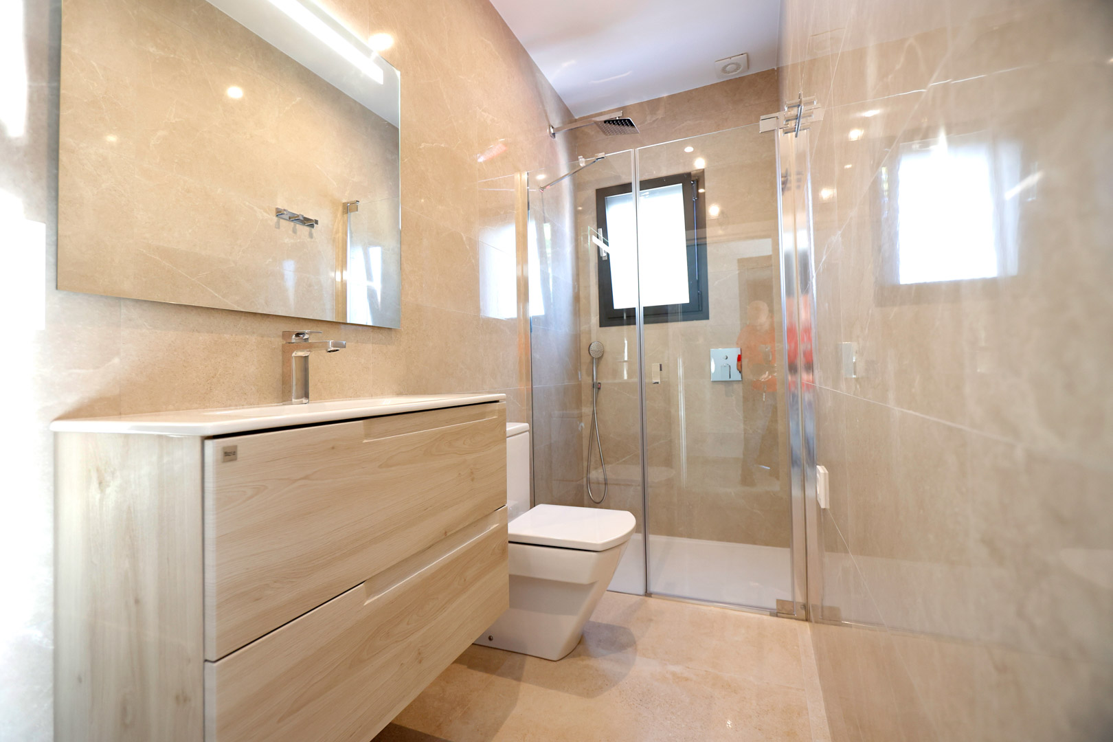 Modernes Badezimmer mit hellem Holzwaschtisch, Glasduschkabine und beigefarbenen Feinsteinzeugfliesen. Mit verchromten Armaturen und integrierter Spiegelbeleuchtung.