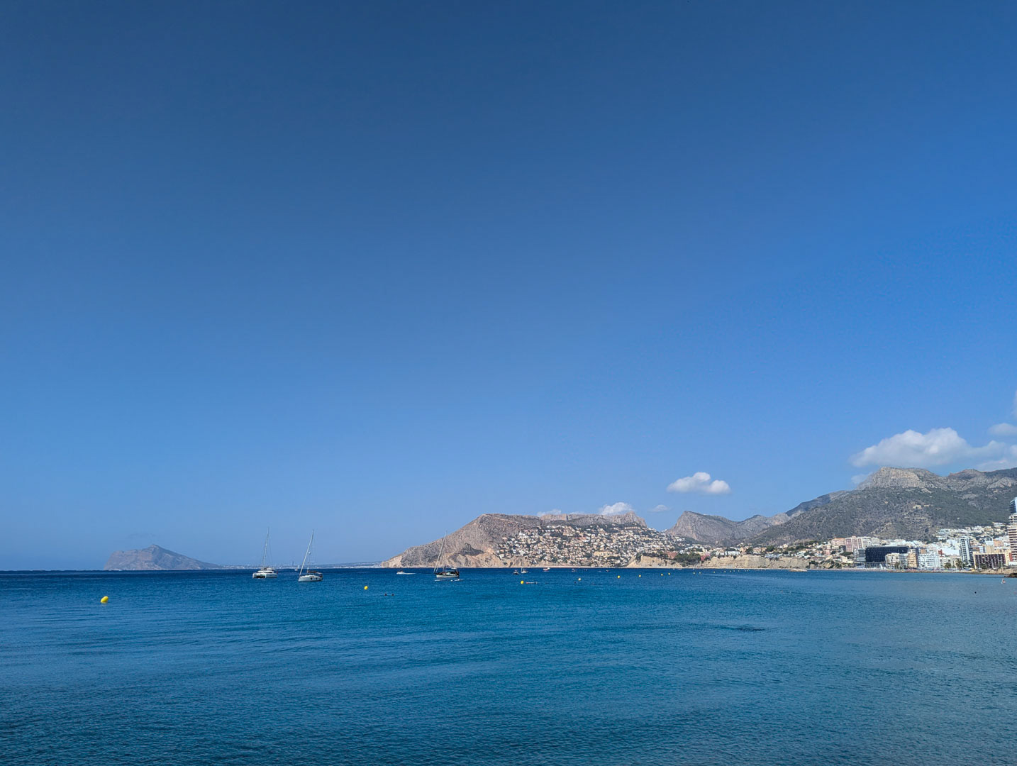 Panoramablick auf das Mittelmeer und die bergige Küste mit vor Anker liegenden Segelbooten unter klarem blauem Himmel.