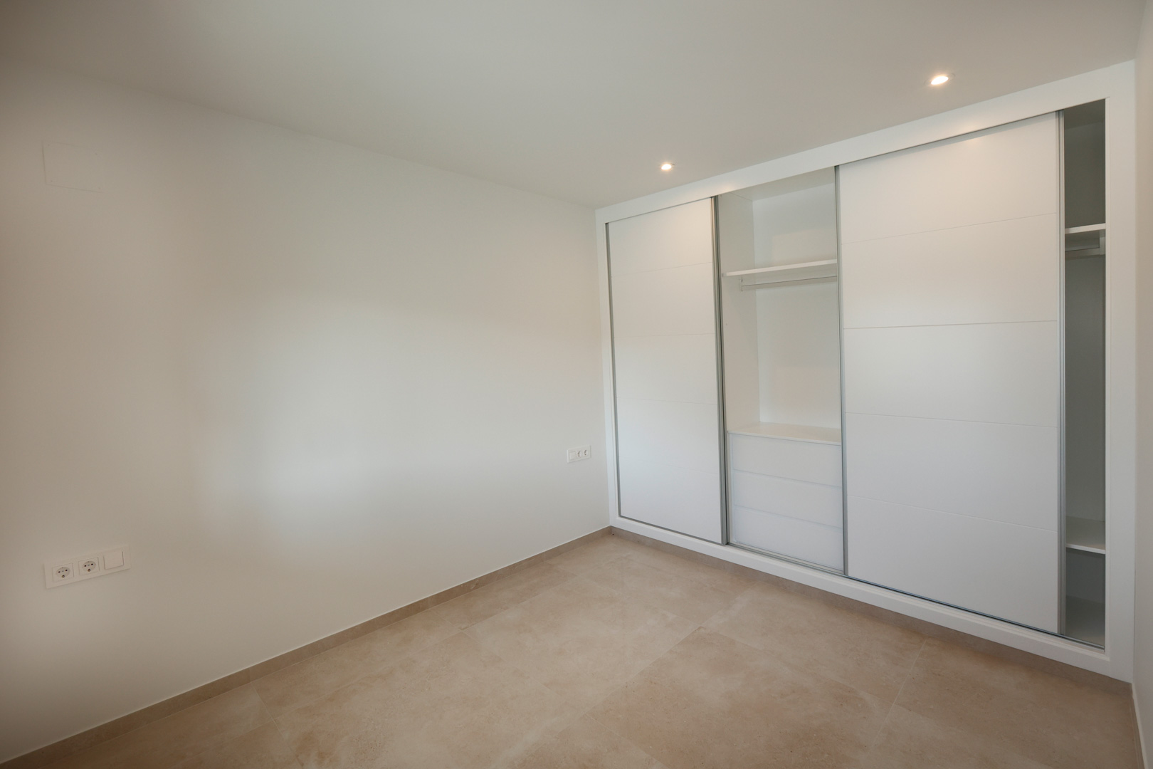 Schlafzimmer mit Einbauschrank mit weiß lackierten Schiebetüren, Feinsteinzeugboden und LED-Einbaustrahlern an der Decke.