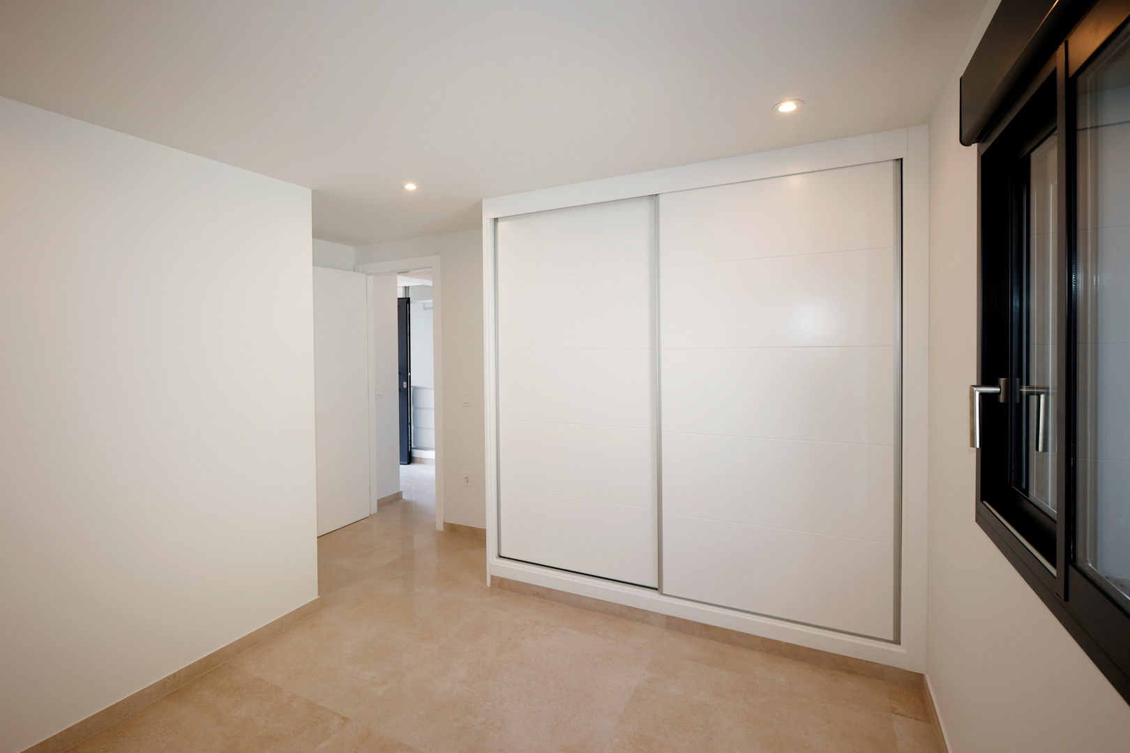 Schlafzimmer mit Einbauschrank mit weißen Schiebetüren, Feinsteinzeugboden und LED-Einbaustrahlern neben einem dunklen Aluminium-Fensterrahmen.