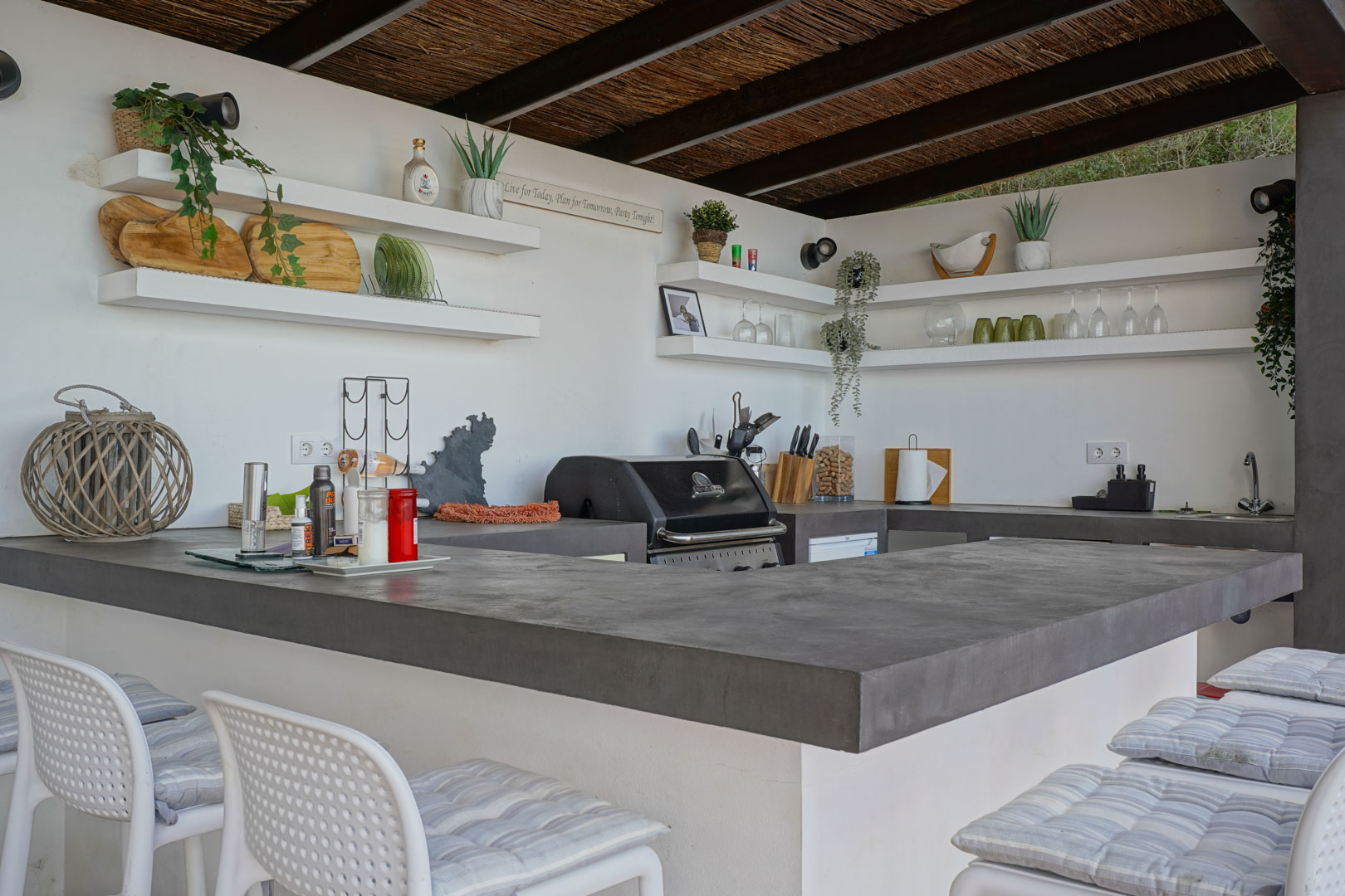 Außen-Sommerküche mit grauen Mikrozement-Arbeitsplatten, integriertem Gasgrill, offenen Regalen und einer Schilfrohrdecke mit dunklen Holzbalken.