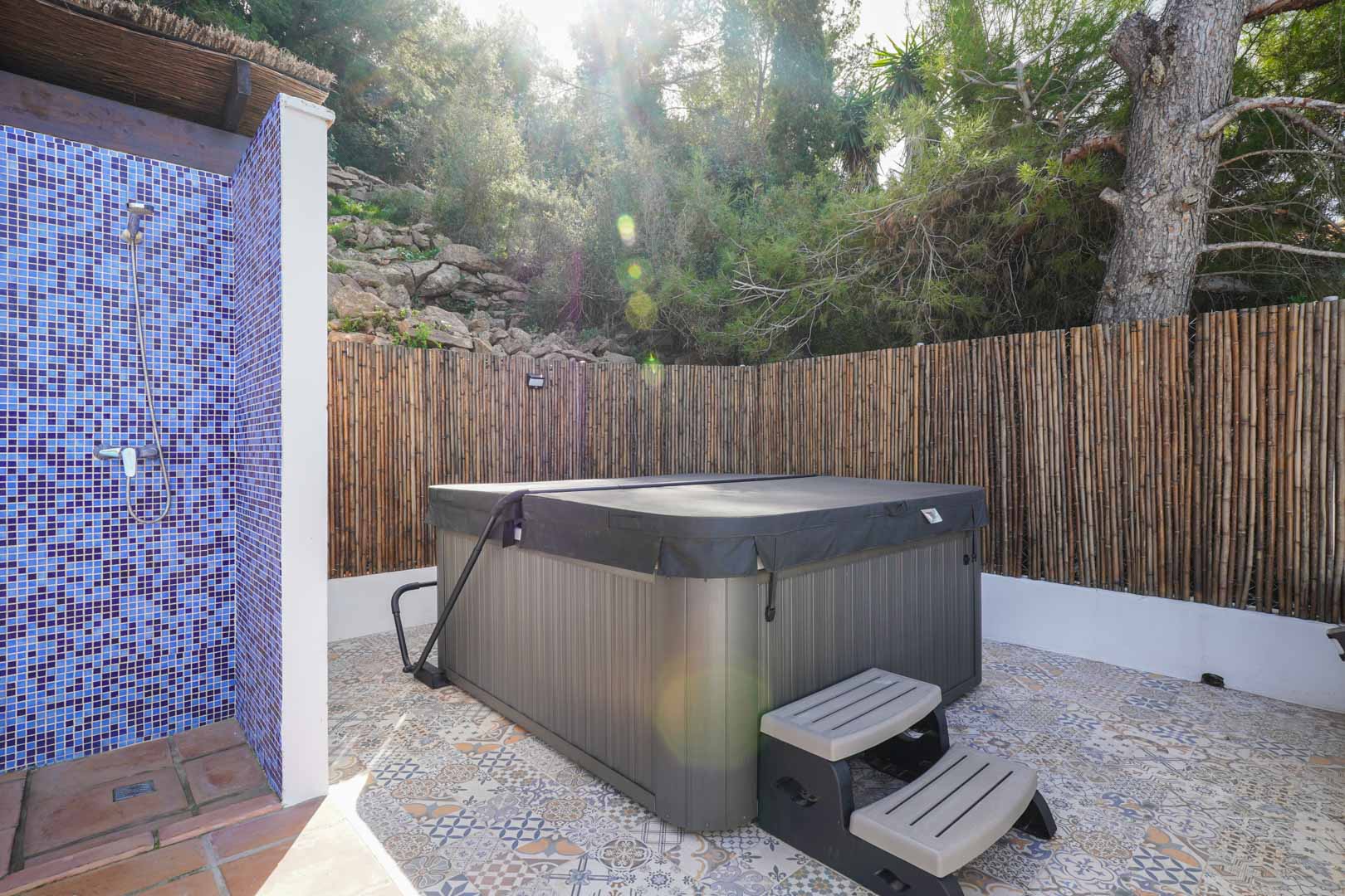 Außen-Wellnessbereich mit abgedecktem Whirlpool, blauer Mosaikdusche und gemustertem Fliesenboden, eingefasst von einem natürlichen Sichtschutz aus Schilfrohr.