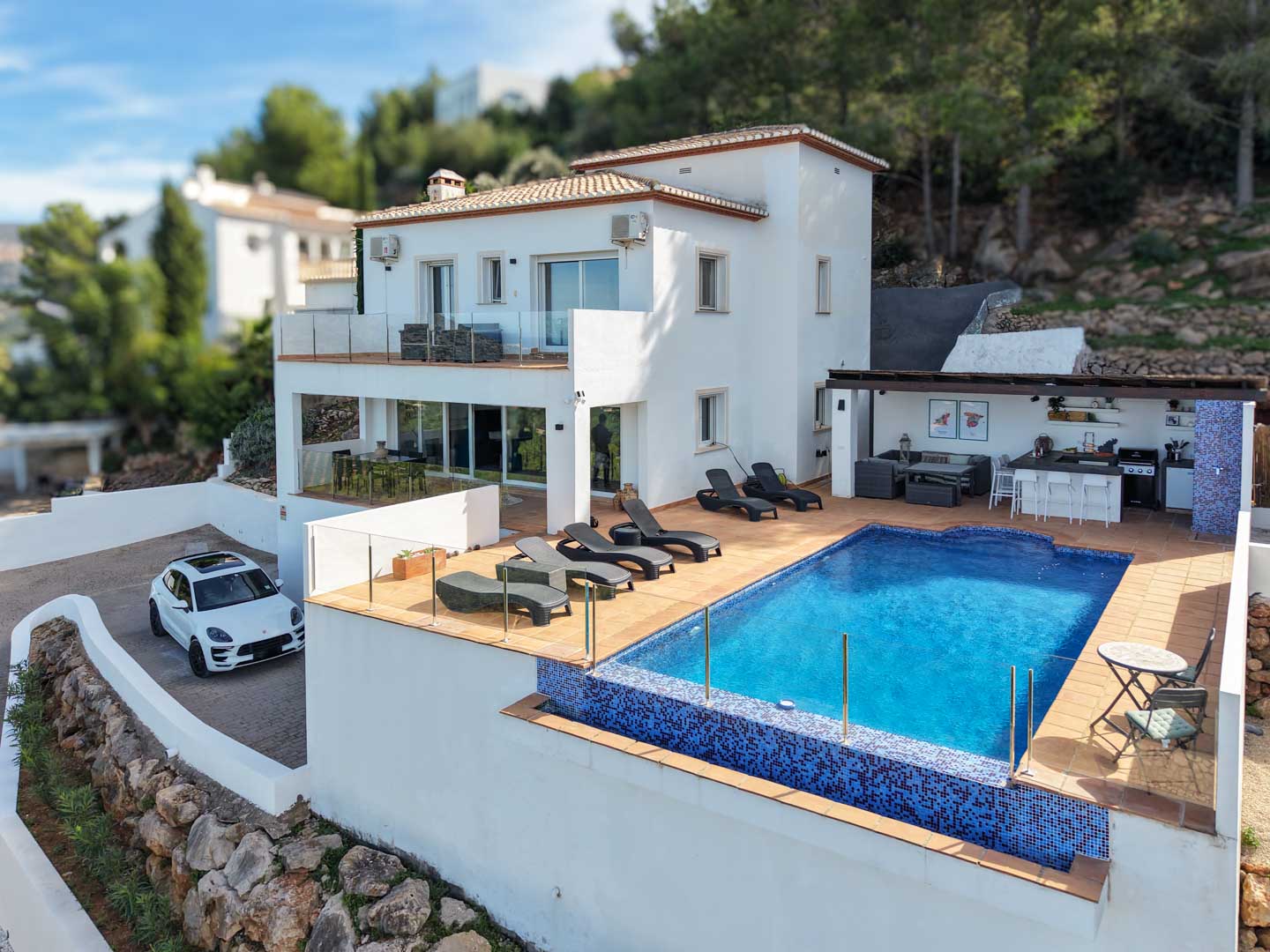 Außenansicht der Villa mit blauem Mosaikpool, Sonnenterrasse, Außenküche unter einer Pergola und weißer mediterraner Fassade in exklusiver Hanglage.