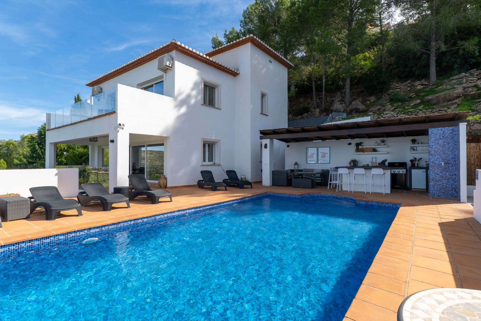 Außenbereich mit Mosaikpool, Terrakotta-Terrasse und überdachter Sommerküche. Die weiße mediterrane Villa liegt direkt an einem bewaldeten Hang mit Pinien.