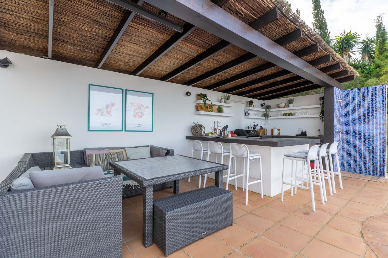 Außenküche und Loungebereich unter einer Holzpergola mit Reetdach, ausgestattet mit einer Bar, weißen Hockern, grauen Polyrattan-Möbeln und Terrakotta-Fliesen.
